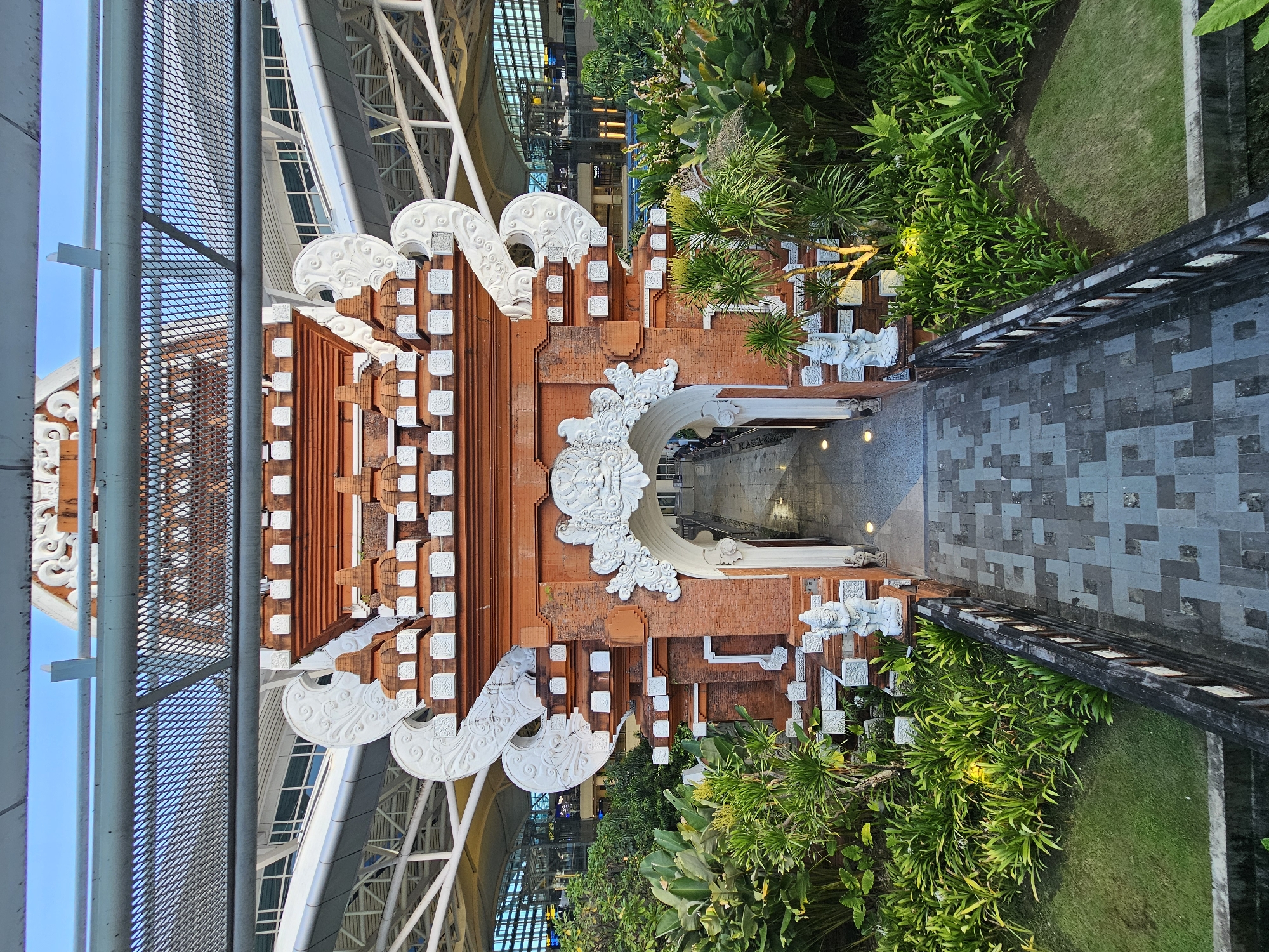 The image depicts a Balinese gate leading to a walkway. The gate is decorated with white carvings on a brick-colored structure. Lush greenery flanks the walkway leading to a dark passage. In the background, modern architecture is visible. The image is a vertical, low-angle shot. The gate is likely located in Kuta, Indonesia, given the style of architecture.