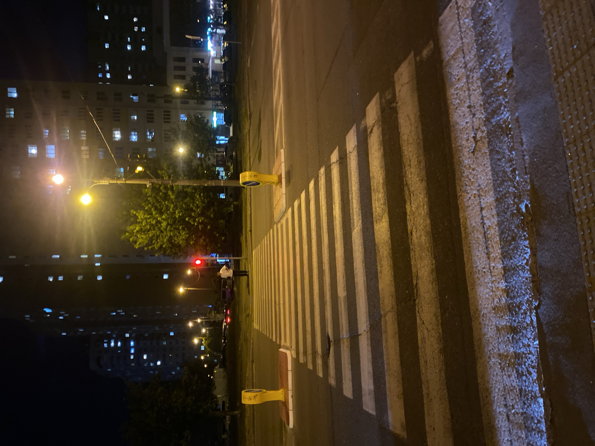 A nighttime street scene in Kyiv, Ukraine, features a pedestrian crossing marked by white stripes on asphalt. Yellow bollards are positioned on either side of the crossing, each with a small red and white base. A street lamp with a bright yellow light illuminates the scene, casting long shadows. In the background, tall apartment buildings with numerous lit windows are visible. A single person, dressed in a white shirt and dark pants, stands on the sidewalk near a red traffic light, facing away from the camera. Cars are visible in the distance, their headlights and taillights creating streaks of light. Trees with green foliage line the side of the road, and streetlights are dotted throughout the urban landscape. The road surface appears wet in places, reflecting the artificial lights.
