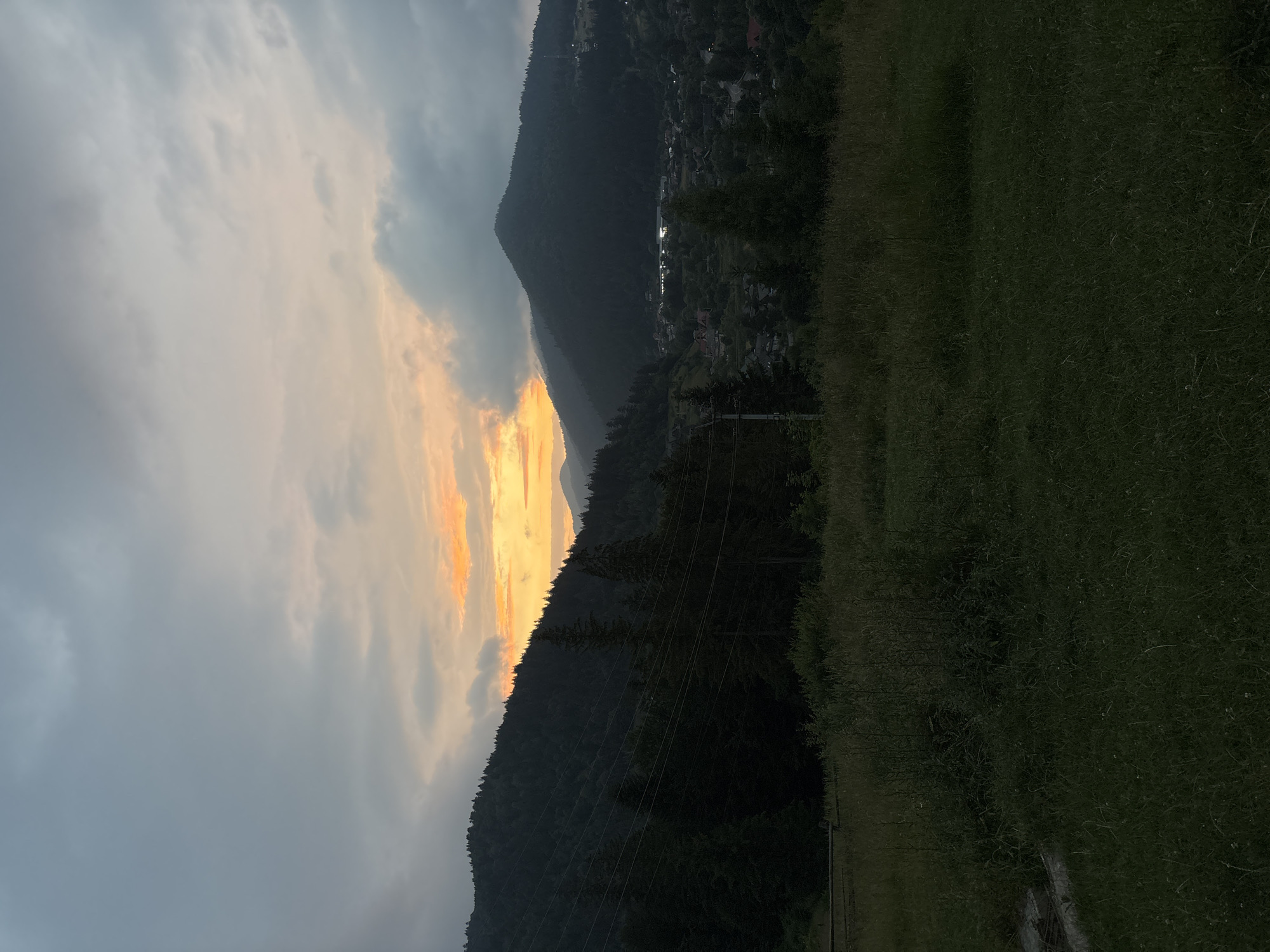 This is a vertical, long shot of a sunset over a mountainous village. The sky is filled with dramatic clouds, with a bright orange and yellow sunset breaking through on the left side of the frame. The sun's rays illuminate the clouds, creating a striking contrast with the darker, more ominous clouds on the left.
In the middle ground, a densely forested mountain slopes down, revealing a village nestled in the valley. The lights of the village are visible, suggesting it's dusk or early evening. The village is surrounded by trees, and a few power lines are visible crossing the frame, indicating human presence and infrastructure.
The foreground is dominated by a grassy field, which appears somewhat wild and untamed. The grass is green and textured, with some patches of dry grass visible. A dark line, possibly a road or path, runs through the grass on the right side of the frame.
The overall mood is serene and dramatic, capturing the beauty of nature as day transitions into night. The time of day is clearly sunset, with the vibrant colors in the sky being the most prominent indicator. The weather appears to be mixed, with both clear and cloudy skies. The location appears to be a rural mountainous area. There is no visible text in the image.