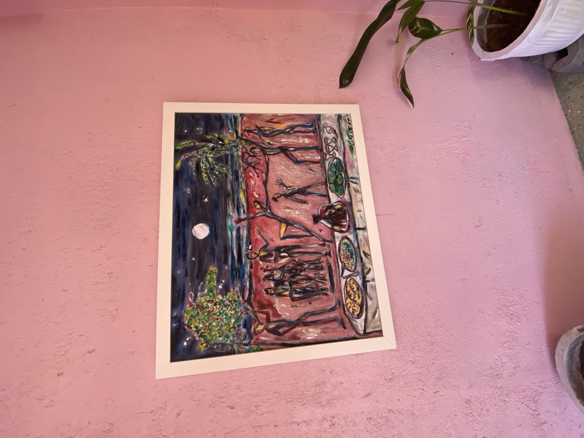 A framed painting hangs on a textured pink wall. The painting depicts a nighttime scene with a moon in the sky. In the foreground, a table is laden with food and drinks. In the middle ground, several figures are gathered, some appearing to be playing music or dancing. In the background, there is a beach or water scene. To the right of the painting, a potted plant with green and white striped leaves is visible. Two concrete planters are partially visible in the bottom left corner. The floor is a speckled gray and green.