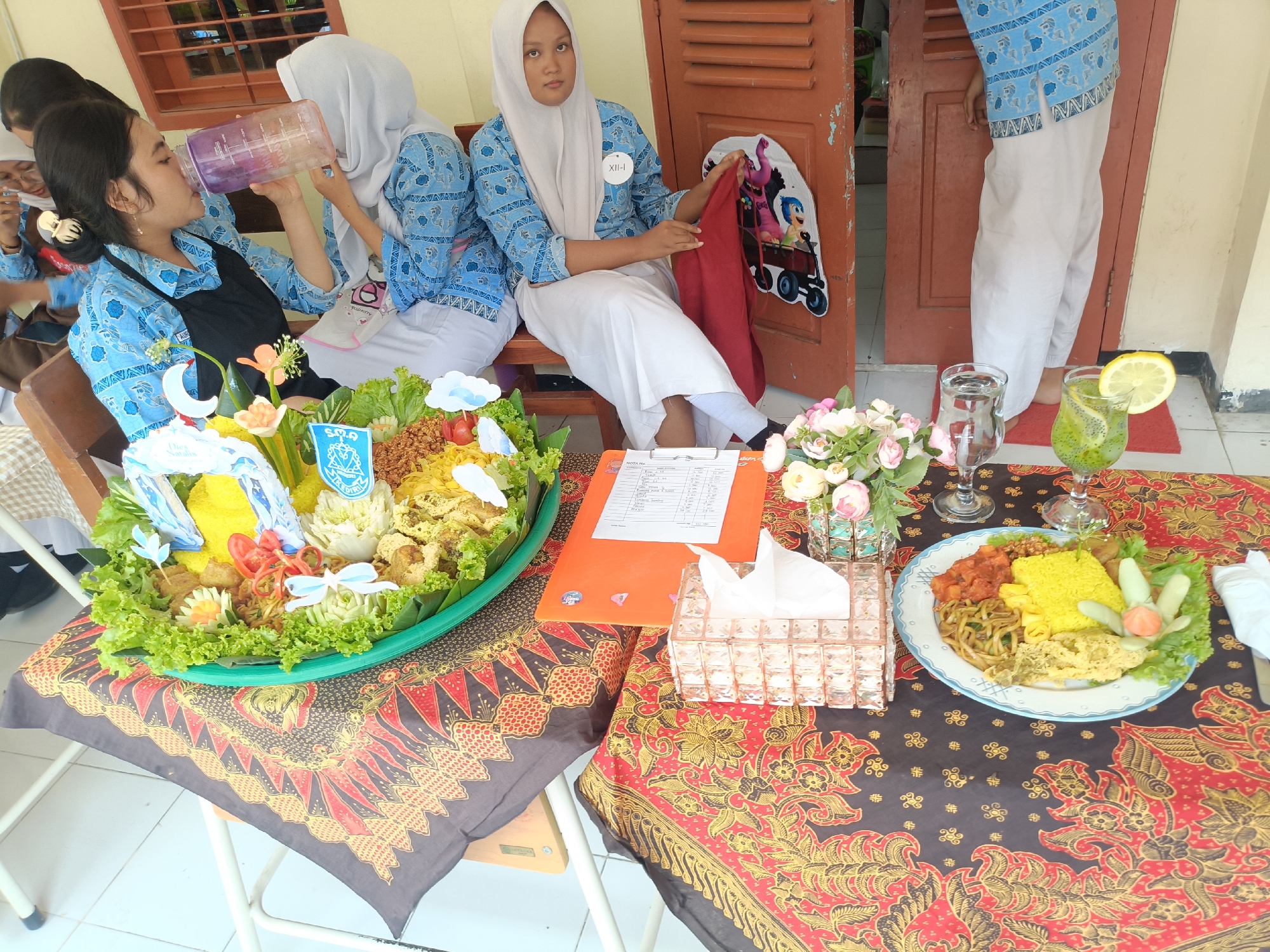 The image depicts a group of young women dressed in traditional blue patterned school uniforms and white headscarves, sitting around a table laden with food. One woman on the left is drinking from a large purple water bottle, while another woman in the center is holding a red cloth. A table in front of them is covered with a vibrantly patterned batik cloth. On the table, there are two large platters of food, one with yellow rice and various side dishes, and another smaller plate with a serving of yellow rice and accompaniments. A bouquet of flowers, a tissue box, a glass of water, and a glass of green drink with a slice of lemon are also visible. A clipboard with papers sits on the table, and a decorative cutout of characters from the movie "Inside Out" is attached to a wooden door in the background. The scene appears to be an outdoor event, possibly a school or community gathering, given the presence of the students and the decorations. The architectural style of the building in the background, with its wooden doors and visible window bars, suggests a location in Indonesia. The batik tablecloth is a strong indicator of Indonesian origin, and the overall atmosphere points to a local cultural event. While the exact location within Kediri City is not explicitly stated, the context suggests a public or institutional setting within the city.