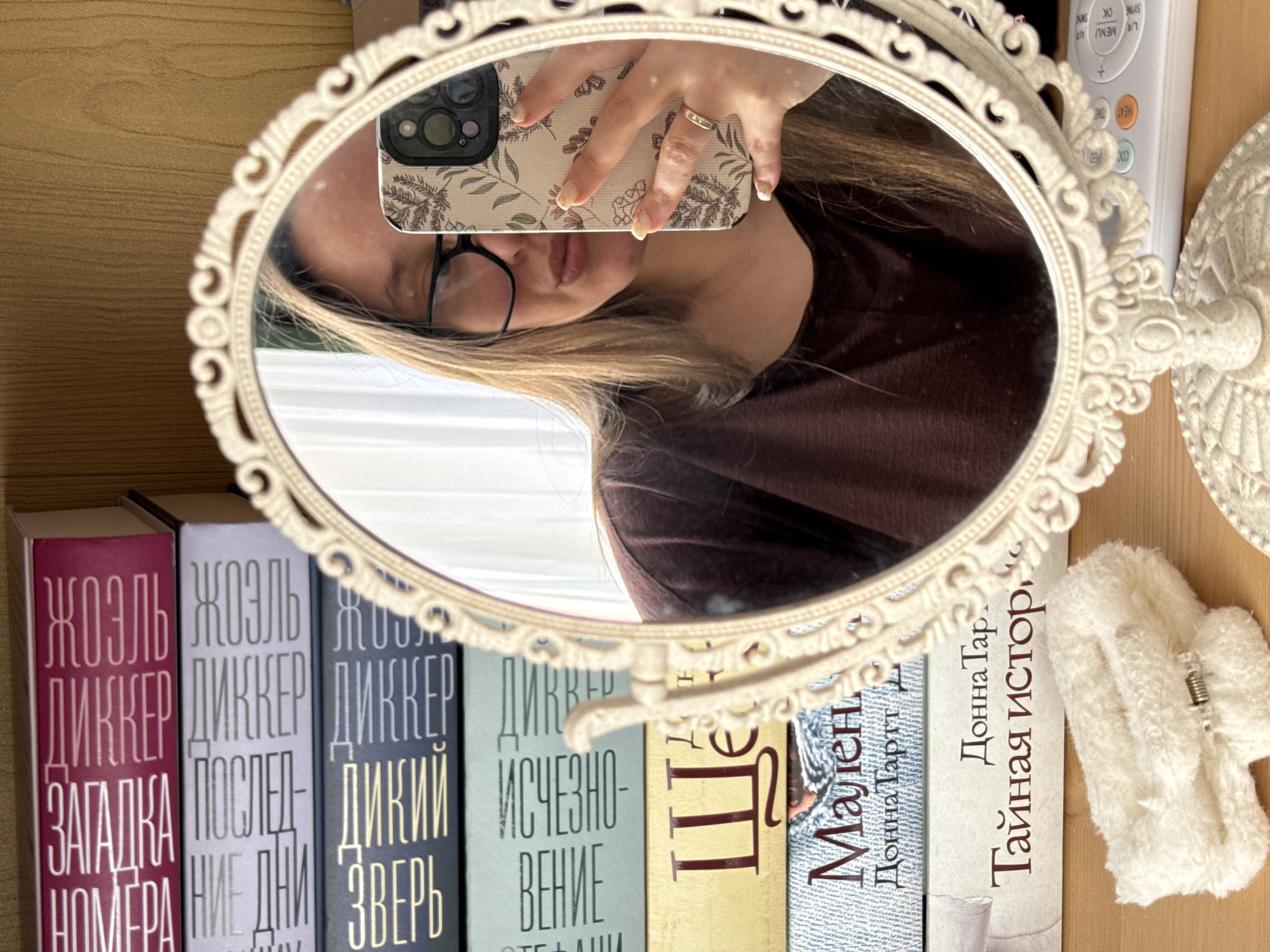 A person reflected in an oval vanity mirror is taking a selfie. The mirror is placed in front of a shelf of books, with titles in Russian. The books appear to be part of the Joel Dicker series and books by Donna Tartt. A white remote control is visible to the right of the mirror. A white hair clip is on the shelf next to the last book. The location appears to be indoors, possibly a home library or bedroom. The city of Krasnodar, Russia, cannot be determined from the image.