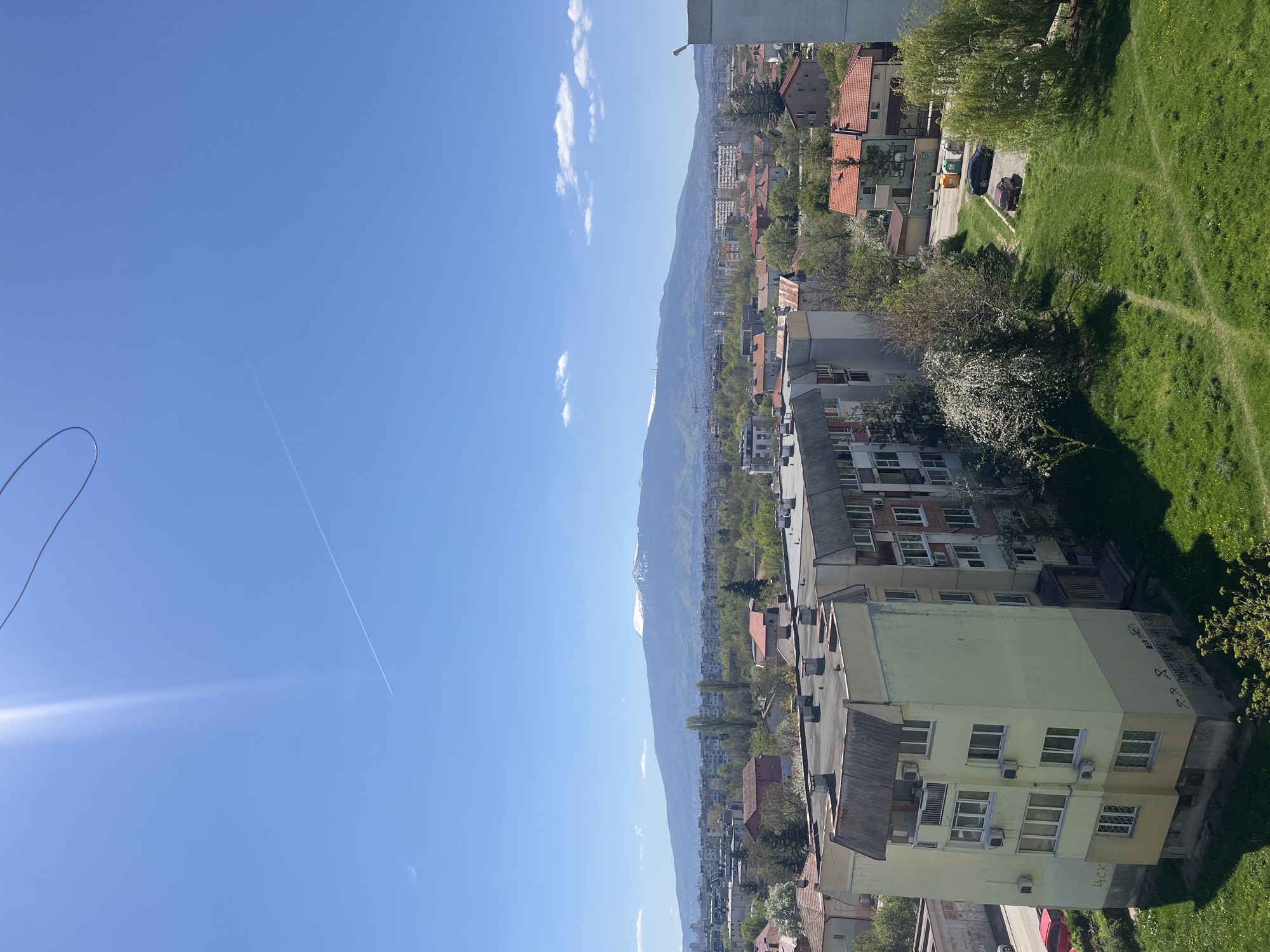 The image shows a cityscape in Sofia, Bulgaria, on a sunny day. A mountain range forms the backdrop. In the foreground are apartment buildings, some with visible graffiti, and patches of green space. The sky is blue with contrails and a few scattered clouds. A section of a roof appears in the top right and a wire is visible in the top left.