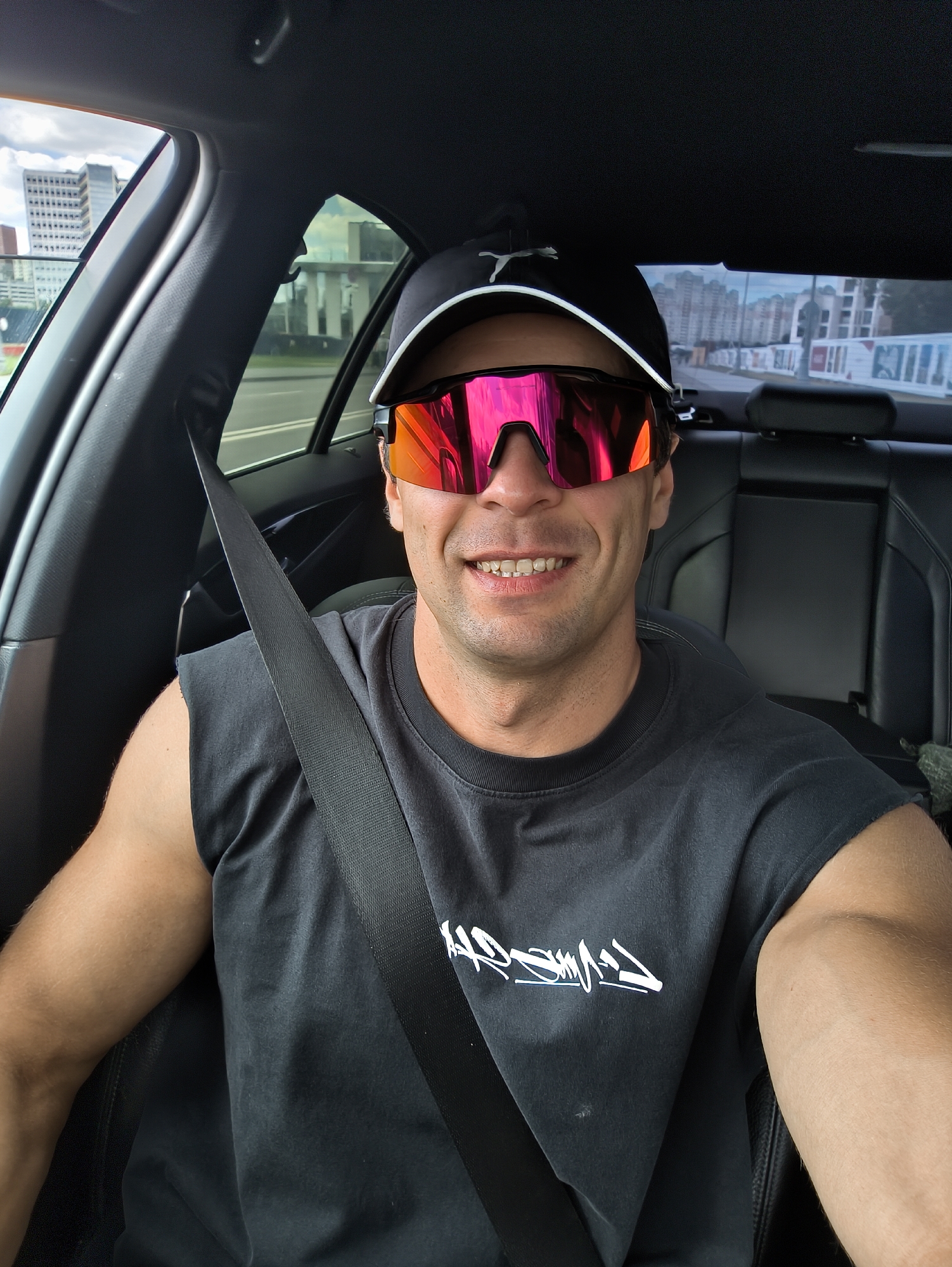 A man in a black Puma baseball cap and black sleeveless t-shirt with a white graphic print is seated in the driver's seat of a car, wearing reflective pink sunglasses. He is smiling, with his mouth open, revealing his teeth. The seatbelt is fastened across his chest. The interior of the car, including black leather seats, is visible. Outside the car windows, city buildings, roads, and a cloudy sky can be seen, suggesting an urban environment, potentially Moscow, Russia.