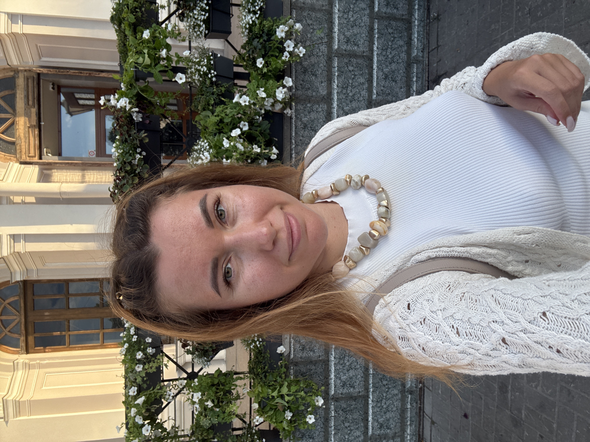A portrait-style photograph features a young woman with long, light brown hair and green eyes, smiling subtly at the camera. She has light freckles across her nose and cheeks. She is dressed in a white, ribbed crew-neck top and a light cream-colored, open-knit cardigan with intricate cable and lace patterns. Around her neck, she wears a necklace composed of large, irregular, light-toned (beige, grey, off-white) stones or beads, interspersed with small gold-colored cylindrical beads. A beige strap, possibly from a bag or backpack, is visible over her right shoulder.
The background on the left side of the frame showcases a building with classic architectural details, including a light-colored facade and an ornate window featuring wooden frames and a rounded arch pattern. Below this, lush green foliage and numerous small white flowers, resembling petunias, spill from window boxes or hanging planters. To the right, behind the woman, a textured grey wall or paved surface is visible. The lighting is soft and warm, suggesting the photo was taken in the late afternoon or early evening. The overall mood is pleasant and casual. No text is visible in the image.