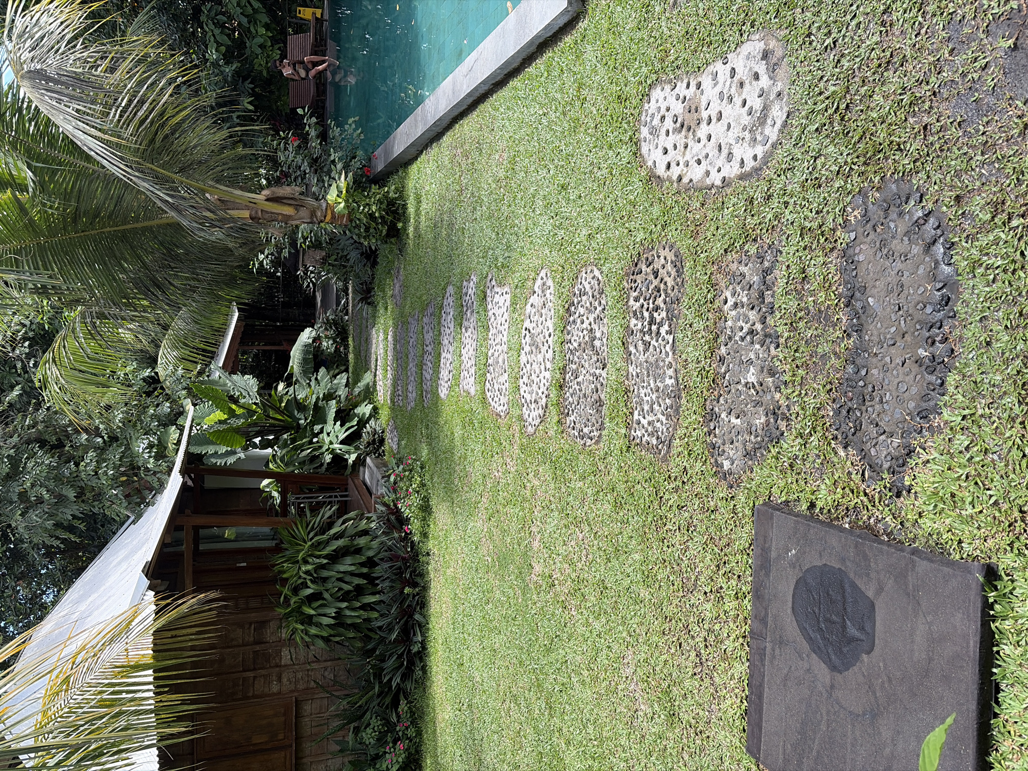 The image depicts a tropical resort setting in Mengwi, Indonesia. In the foreground, a pathway of irregularly shaped stepping stones made of concrete and embedded pebbles winds through a manicured lawn. To the left, a rustic wooden bungalow with a corrugated metal roof is partially visible, surrounded by lush green foliage including large-leafed plants and palm fronds. To the right, a bright turquoise swimming pool with a concrete edge takes up a significant portion of the frame. In the background, a woman is seated on a wooden bench by the pool, holding a phone. The environment suggests a peaceful and relaxing atmosphere, typical of a tropical vacation destination.