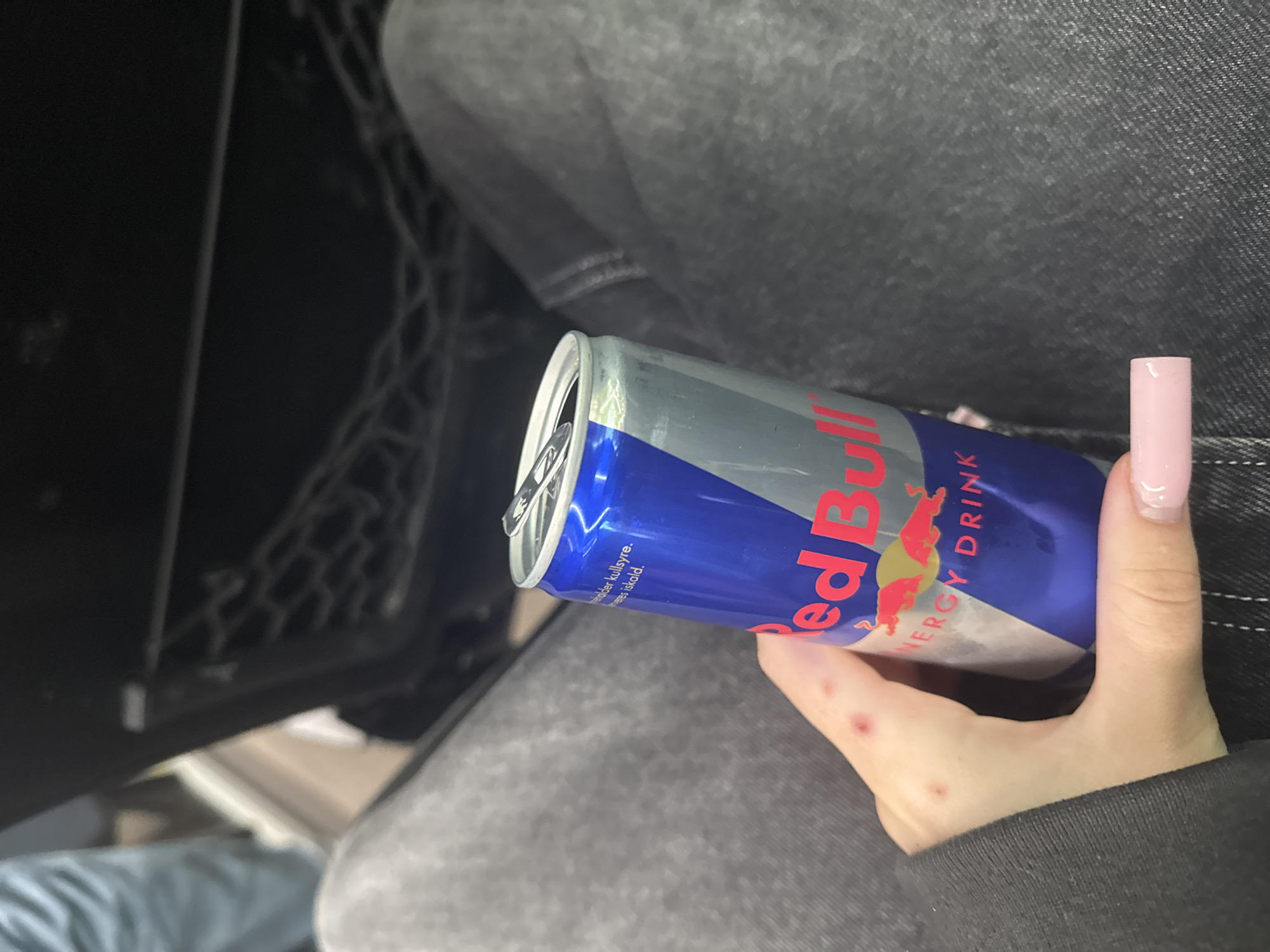 In Skatval, Norway, a person's hand, adorned with pink nail polish and showing red marks on the knuckles, holds a partially opened Red Bull energy drink can. The can, featuring the brand's iconic blue and silver design, is positioned against a backdrop of dark grey denim-like fabric, suggesting the person is wearing jeans. The background also includes a dark, possibly black, mesh pocket and a hint of blue fabric. The lighting is dim, creating shadows and emphasizing the texture of the clothing.