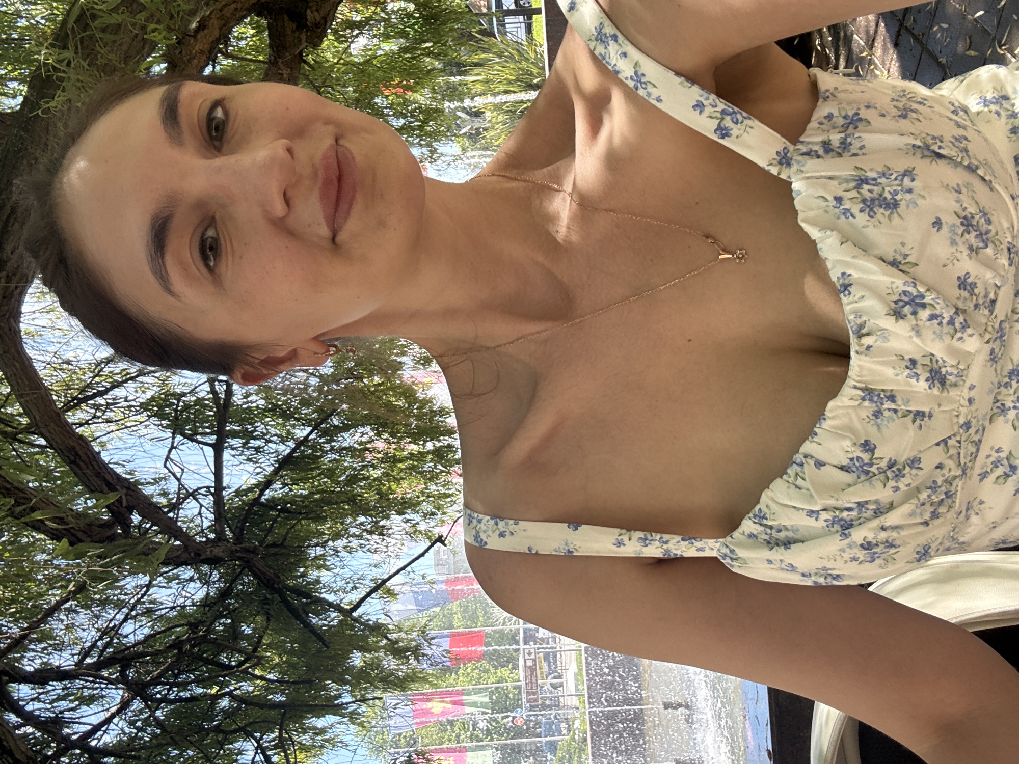 A young woman is captured in a bright, outdoor portrait on a sunny day. The image is oriented vertically, showing her from the upper chest up, with her head slightly tilted. She has dark hair pulled back, distinct eyebrows, and is looking directly at the camera with a gentle, understated smile. She is wearing a light-colored dress with delicate blue floral patterns and thin shoulder straps. A fine gold-colored chain necklace with a small, ornate pendant, possibly a flower, is visible around her neck.
The setting appears to be a verdant park or garden. To the left of the woman, the thick canopy and branches of a large tree with lush green leaves are prominent, with patches of clear blue sky visible through the foliage. To her right, a sparkling water feature, possibly a fountain or pool, can be seen with visible water splashes. In the further background beyond the water, there are several colorful flags (including red, green, and yellow) and hints of distant urban structures. The scene is well-lit, suggesting either late morning or early afternoon. The overall mood is serene and pleasant. No discernible text is visible in the image. The environment is consistent with a vibrant outdoor location like a park in Sochi, Russia.