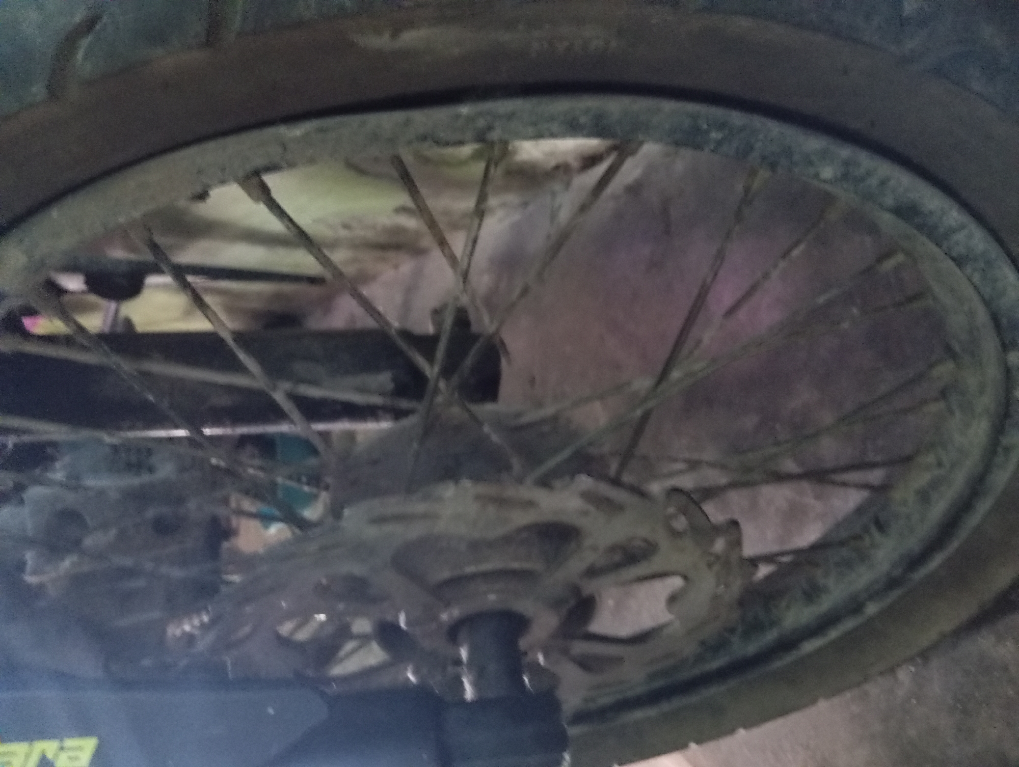 A close-up, slightly blurred image shows the rear wheel of a motorcycle parked on a concrete surface. The motorcycle wheel has a disc brake and spokes. The background is indistinct but appears to be an outdoor setting, possibly a street or alleyway. The overall lighting suggests it is daytime. The image does not provide specific visual cues to pinpoint its location to Rejosari, Indonesia.