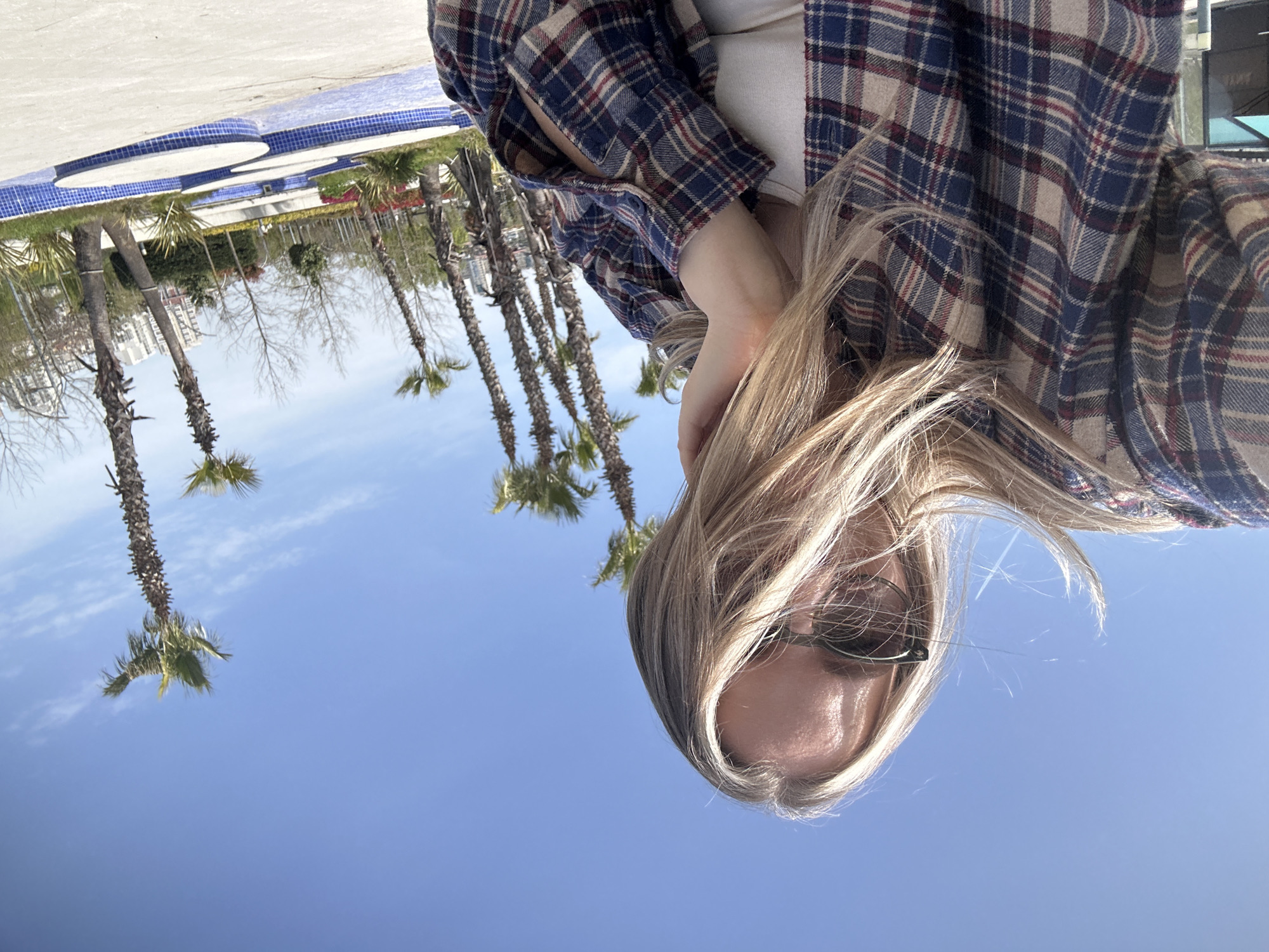A person is looking upwards towards the camera, wearing sunglasses and a plaid shirt. The location appears to be a park in Başakşehir, Turkey, characterized by palm trees and a bright blue sky. In the background, there is a water feature with blue and white tiles and some buildings.