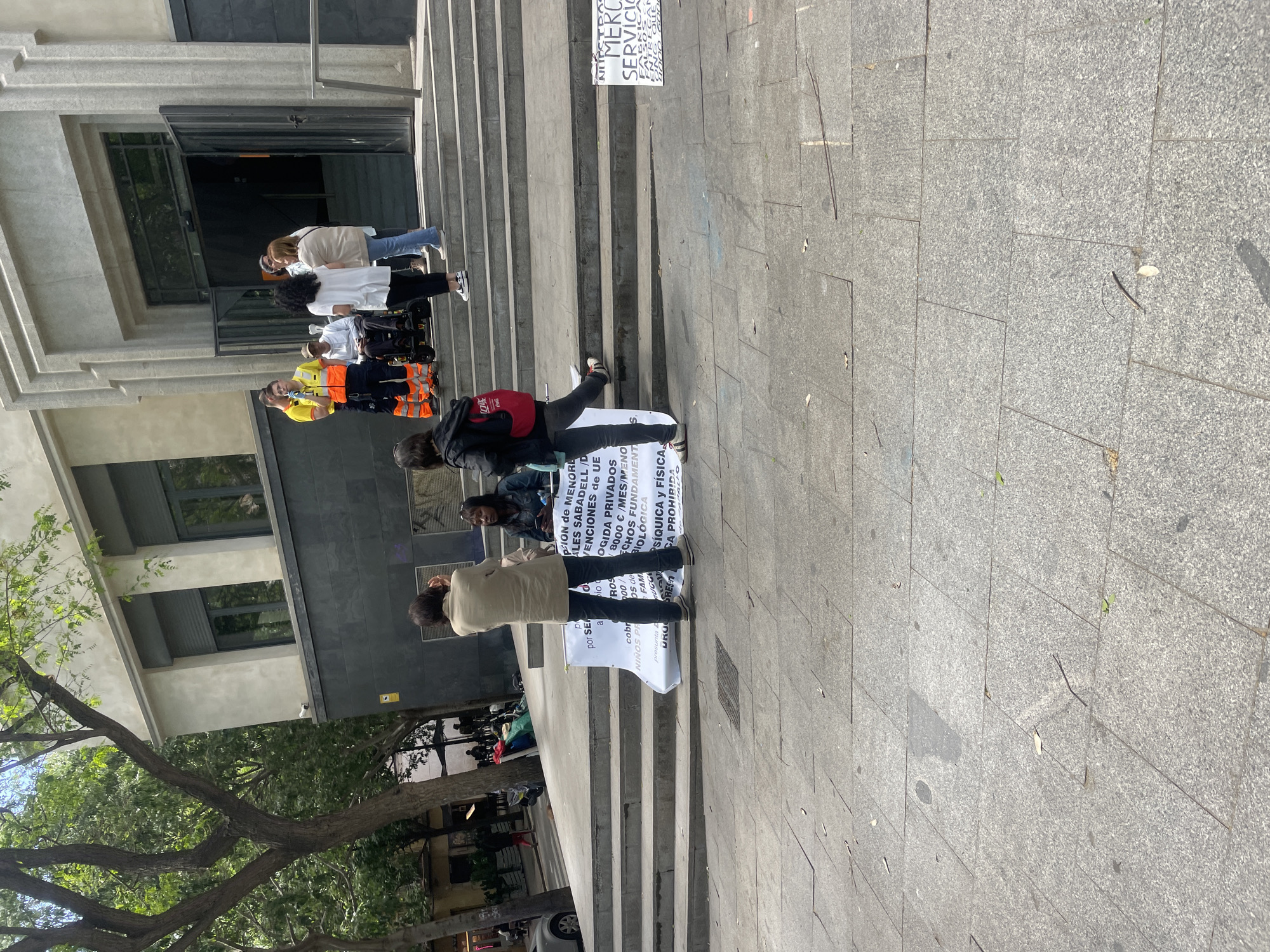 Here's a detailed description of the image:
**Scene:** An outdoor scene on a sunny day, likely in an urban area. The primary focus is a group of people standing on the steps of a building, possibly a government building or public institution. The foreground consists of a paved area, likely a sidewalk or plaza. A large tree with green leaves is visible on the left.
**People:**
* A group of people is standing on the steps, some holding signs.
* Some of them are wearing casual clothing, others are wearing safety vests.
* One person is in a wheelchair.
* The protesters are likely positioned in front of the building to gain attention.
**Objects:**
* Several signs are being held by the people, containing text in Spanish.
* A building with a dark-colored facade, windows, and a large door.
* Steps leading up to the building.
* A red backpack is worn by one of the people.
* A tree with full foliage on the left side of the frame.
**Actions:**
* The people are standing and holding signs, likely protesting or demonstrating.
* Some may be engaged in conversation or chanting.
**Environment:** The scene appears to be in a public area, likely in a city. The lighting suggests a clear day. The architecture is modern and possibly institutional. There are visible plants and greenery which indicates the area may have public parks or gardens.
**Location Specific Inference:**
Given that the image includes the Spanish text "Sabadell", a town in Catalonia (near Barcelona, Spain), it is likely that the location of the image is in Barcelona, Spain or a nearby city in Catalonia.