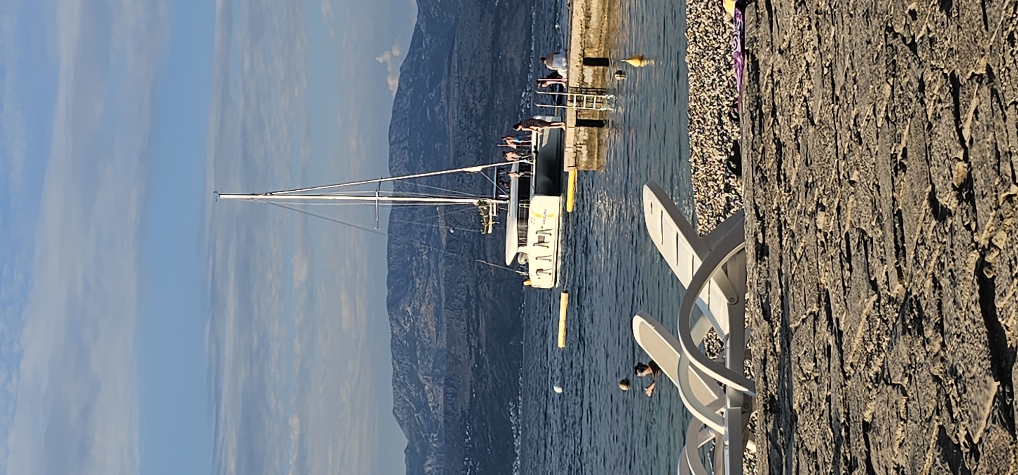 A wide-angle, vertical photograph captures a scenic view of a harbor on a sunny day with a few clouds in the sky. In the foreground, two white lounge chairs are positioned on a pebble beach. Beyond the chairs, the clear, calm water of the sea extends to a dock where several people are gathered. A white catamaran, with a tall mast and sails furled, is moored to the dock. In the background, a mountain range with sparse vegetation overlooks the coastline. The overall scene depicts a tranquil day at a coastal location in Supetar, Croatia.