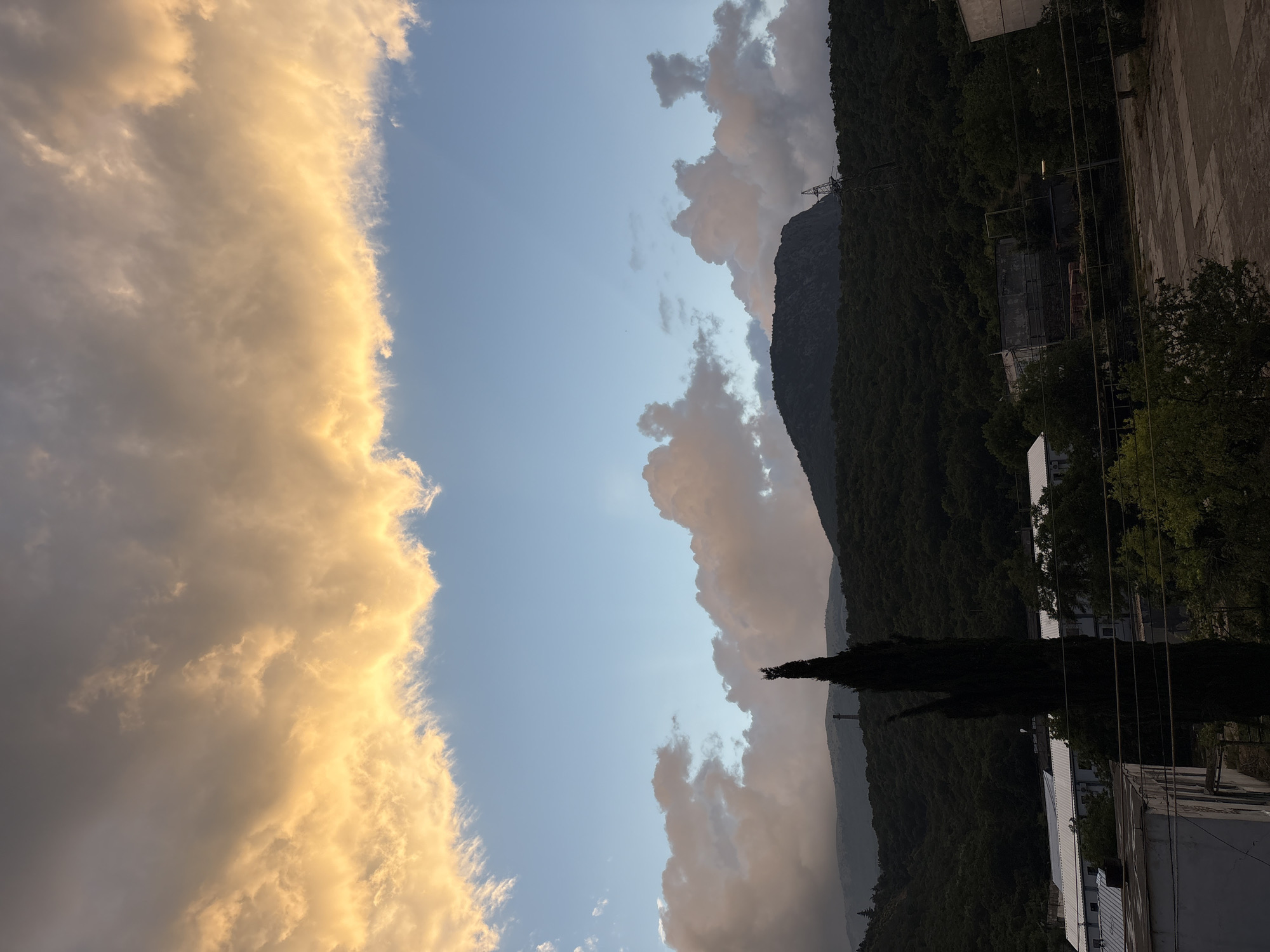 A wide-angle, outdoor, vertical shot shows a dramatic sunset over Malyi Mayak, Ukraine. The upper portion of the frame is dominated by a band of luminous, orange-gold clouds, catching the last rays of sunlight. Below this, the sky transitions to a soft blue, dotted with scattered, pinkish-tan clouds. The middle ground features the dark, tree-covered slopes of mountains, with a prominent, dark, needle-shaped evergreen tree in the foreground, silhouetted against the sky and the distant landscape. Some structures, appearing to be buildings or industrial facilities, are partially visible among the trees and at the base of the mountains. The lower foreground includes a paved area and the rooftops of buildings, with some power lines crisscrossing the scene. The overall impression is one of natural beauty contrasted with human settlement.