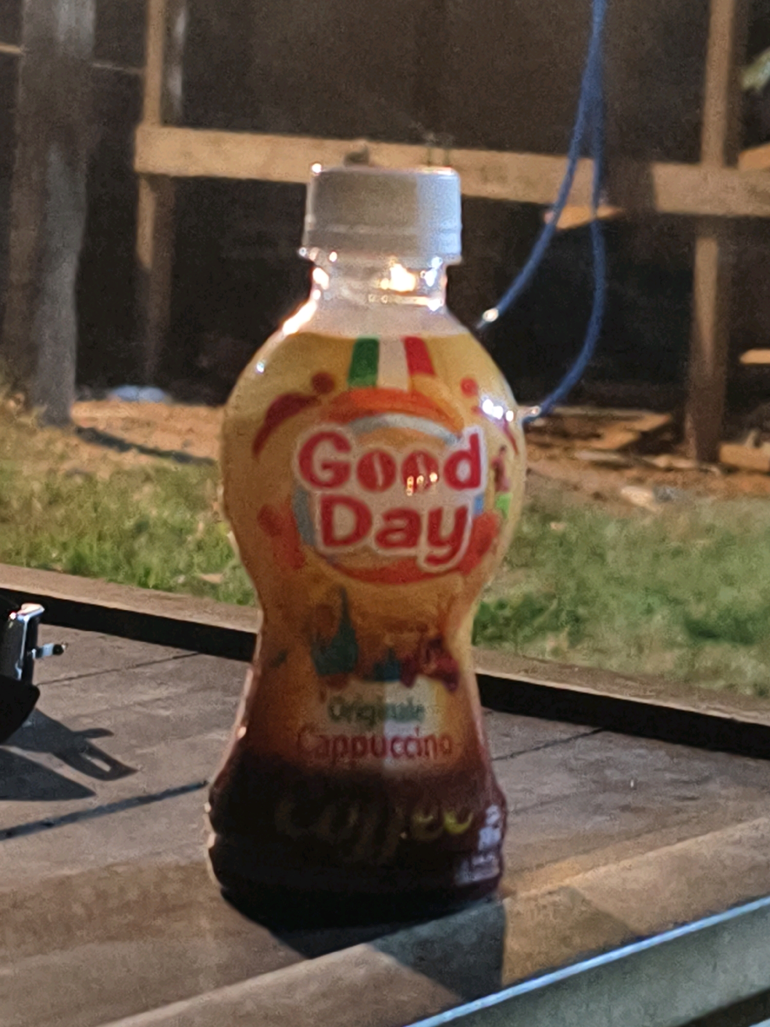 A clear plastic bottle containing a brown liquid with a white cap is positioned on a dark wooden surface. The bottle has a yellow label with red lettering that reads "Good Day Cappuccino". The background is blurred and appears to be an outdoor setting with grass and wooden structures. There are no people or animals visible in the image. The lighting suggests it is either dusk or dawn, or an artificially lit outdoor area. The scene does not provide any specific indicators of its location in Muara Bulian, Indonesia.