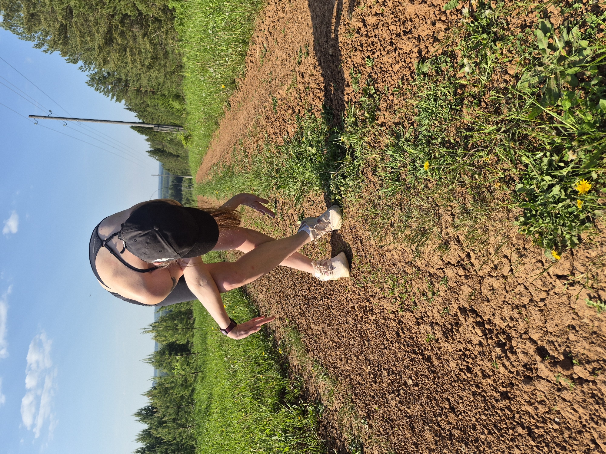 A woman is standing on a dirt road surrounded by green grass and trees under a bright blue sky. She is wearing a black tank top, black shorts, white shoes, and a black baseball cap. She is bent over, with her hands reaching towards the ground. The road extends into the distance, lined with trees and power lines.