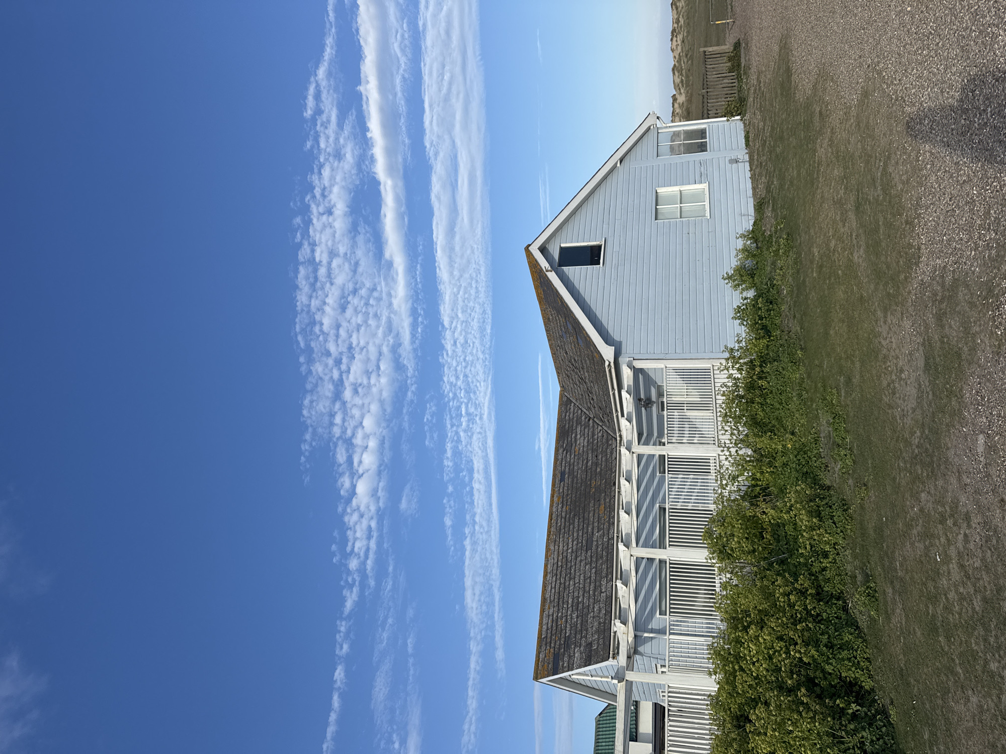 The image shows a light blue, two-story house with a dark grey roof in East Suffolk, United Kingdom. The house has a white balcony railing. The sky is blue with long, thin cloud formations. Ground cover vegetation and gravel surrounds the house. A fence is visible in the background.