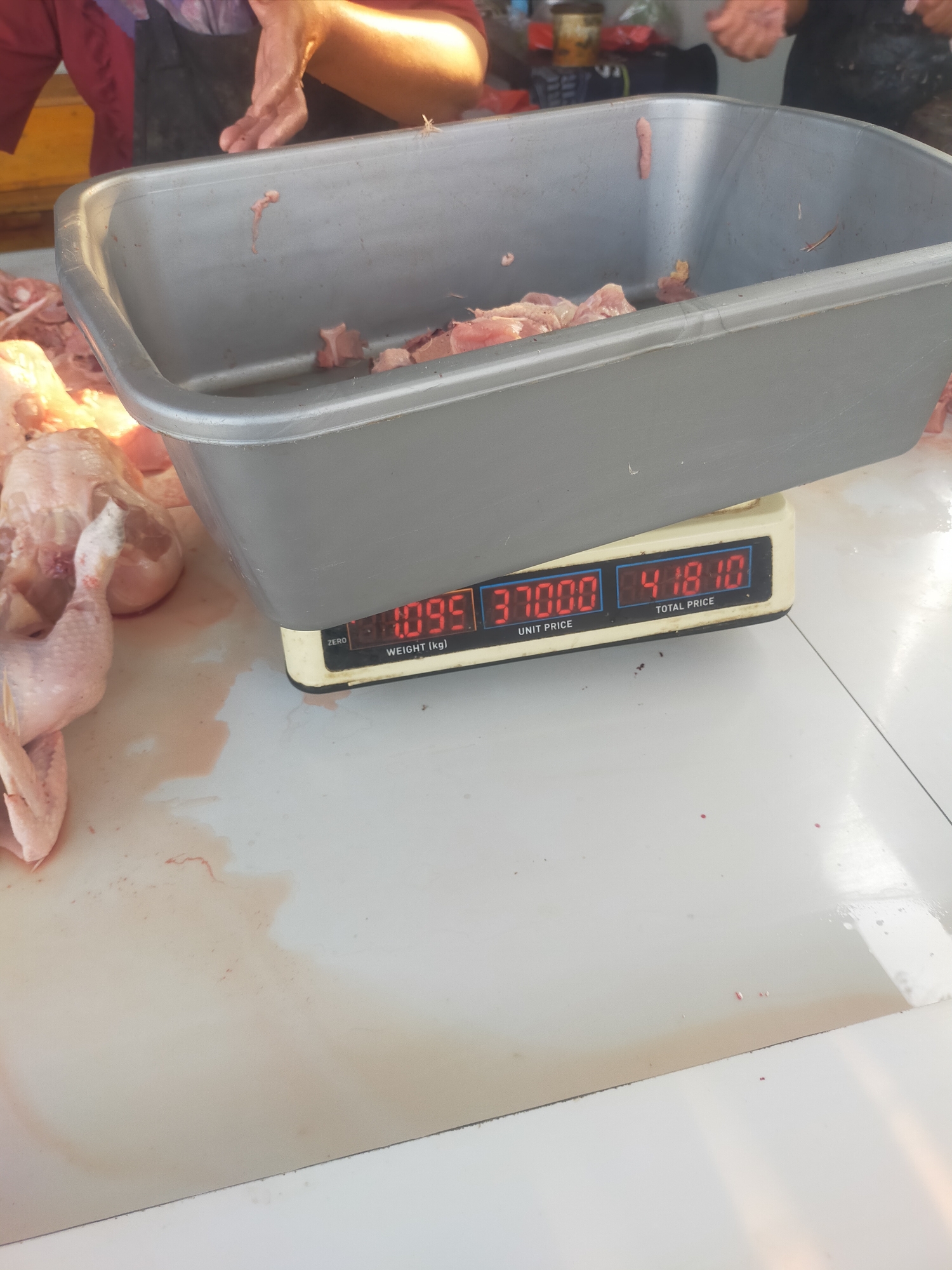 The image shows a close-up view of a digital scale placed on a white surface, likely a table. The scale displays three values: "1.095" under "WEIGHT (kg)", "37000" under "UNIT PRICE", and "418.10" under "TOTAL PRICE". To the left of the scale, there are several raw chicken pieces, some of which are in a gray plastic tub. The chicken appears to be partially processed, with some parts intact and others cut. The white surface is stained with red liquid, likely blood, and there are also a few scattered droplets of red liquid on the surface further away from the chicken. In the background, blurred figures of people can be seen, one of whom is wearing a red garment. The lighting suggests an indoor setting, possibly a market or a food processing area. The scene appears to be in Tegalandong, Indonesia, given the context provided.