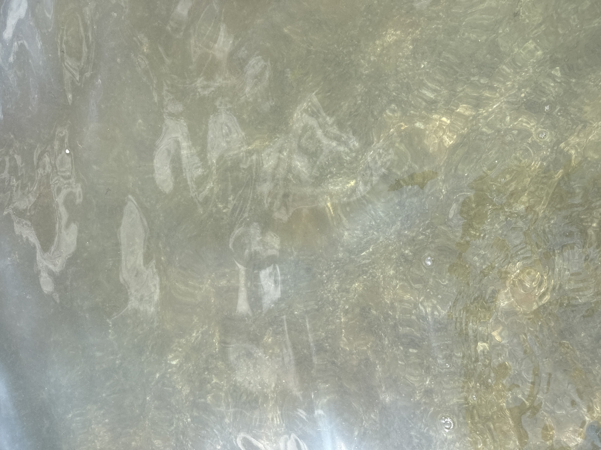 The image displays a shallow body of water, possibly a puddle or a small section of a larger body of water like the Caspian Sea in Aktau, Kazakhstan. The water is murky and reflects the sky and possibly the ground beneath. There are subtle ripples on the surface, indicating some movement. Light glints off the water in certain areas, creating a shimmering effect. The overall impression is one of an aquatic environment with natural light interacting with the surface of the water. There are no visible people, animals, or manufactured objects in the frame.