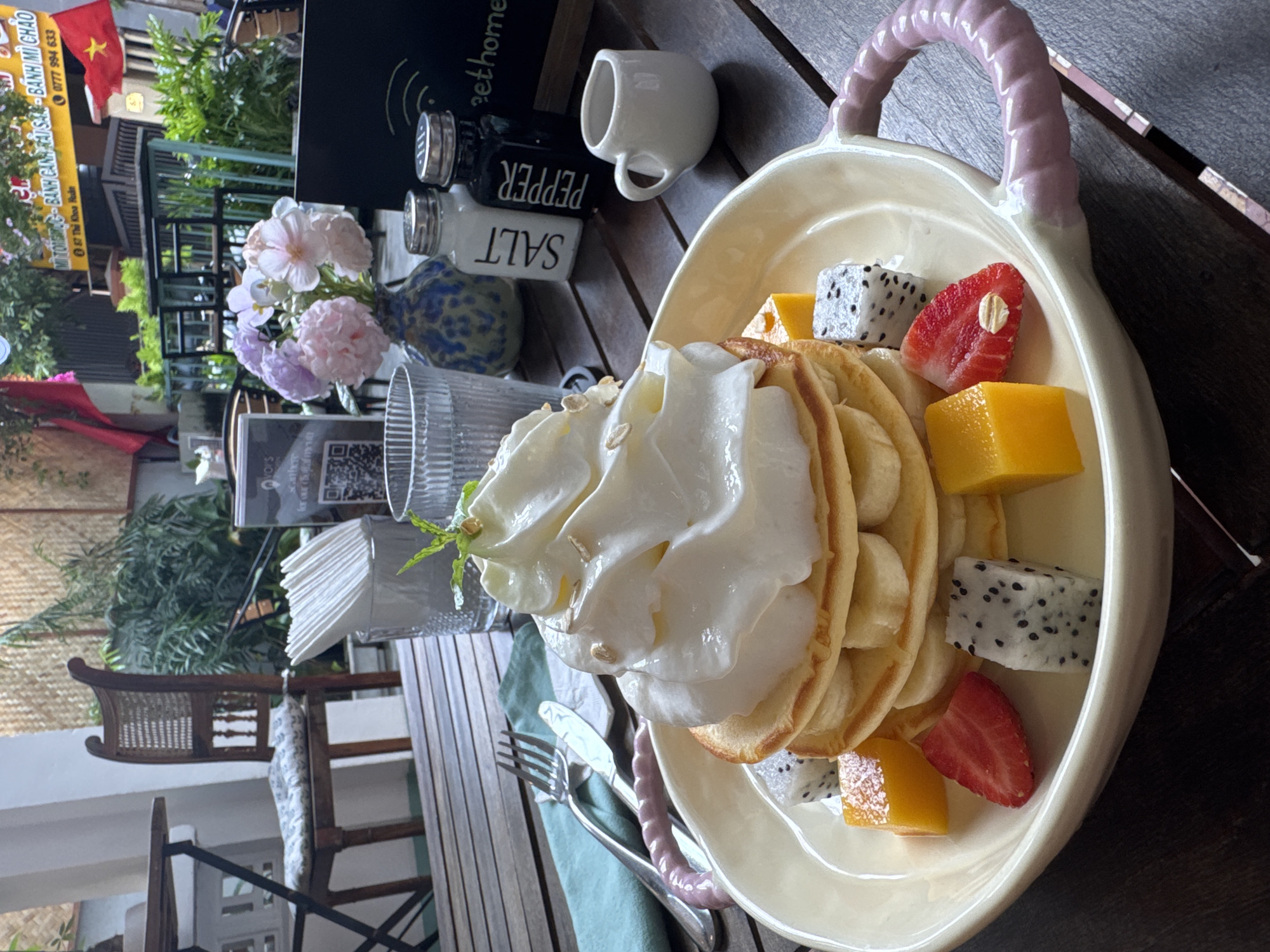 A still, eye-level, full shot captures a table setting featuring a plate of pancakes laden with whipped cream and assorted fruits, including mango, strawberry, and dragon fruit. The plate is positioned on a wooden table, complemented by cutlery wrapped in a light green napkin. Adjacent to the plate, are condiments such as salt and pepper shakers, a small white cup, and a vase holding pink flowers.
In the background, a menu card stands alongside a glass. Further behind, indoor plants are visible, along with a glimpse of a partially obscured Vietnamese flag, situated near the signage of a local business. The sign advertises dishes like "BANH MI CHAO" and "BANH CANH CAI SAN," and includes the phone number "0777 994 633" and the address "67 Thi Khoa Huan."
The scene suggests an outdoor dining area, possibly a café, in Đà Nẵng, Vietnam, as indicated by the Vietnamese signage and flag. The setting implies a casual dining experience with a focus on local cuisine.