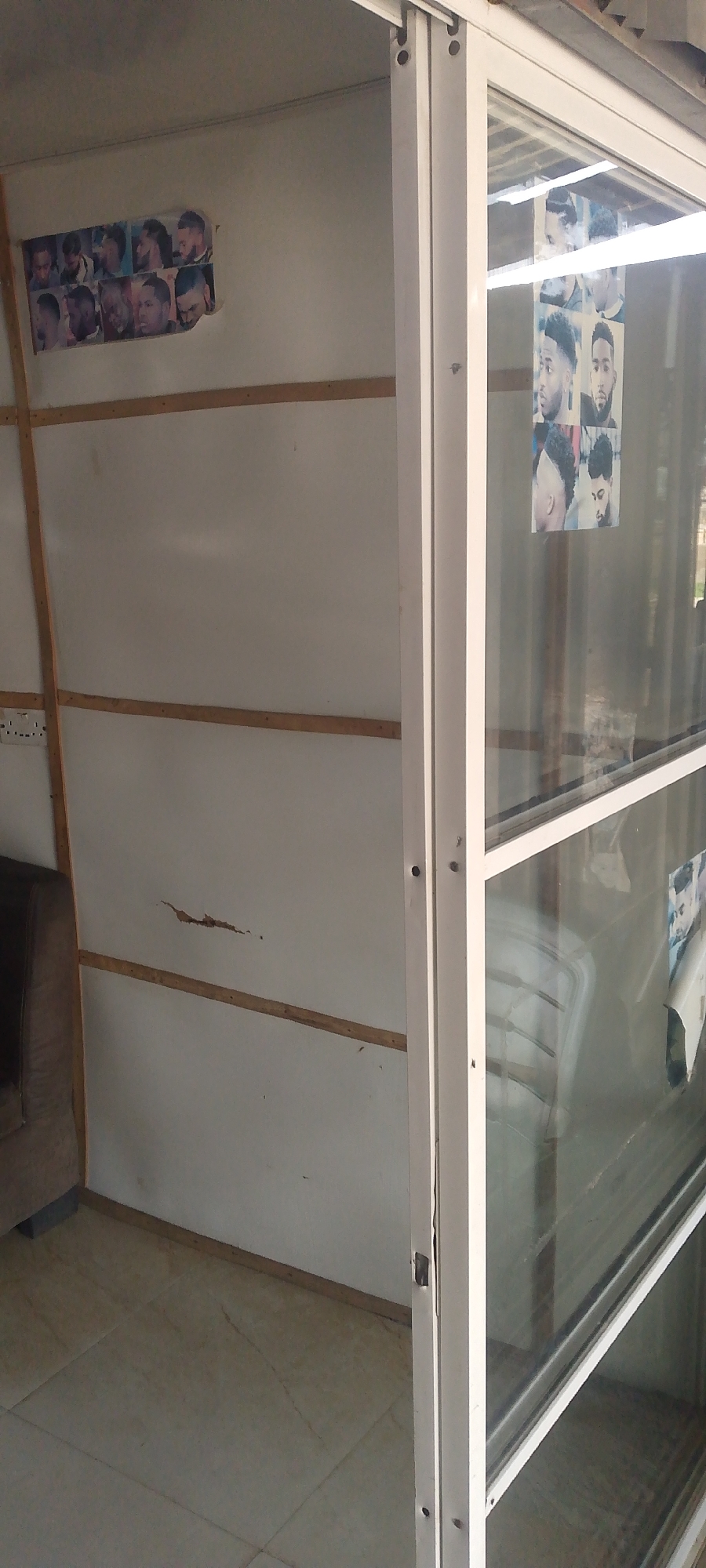 The image shows the interior of what appears to be a small booth or room, possibly a barbershop, located in Jalingo, Nigeria. The walls are white, with horizontal wooden slats for support. On the left wall, there is a collage of images of men's hairstyles. To the right, a large glass enclosure is visible, revealing a display of more hairstyle images on the inside. The floor is tiled. A portion of a brown couch is visible in the lower left corner. The overall impression is one of a commercial establishment, likely a barbershop, in a developing urban setting.