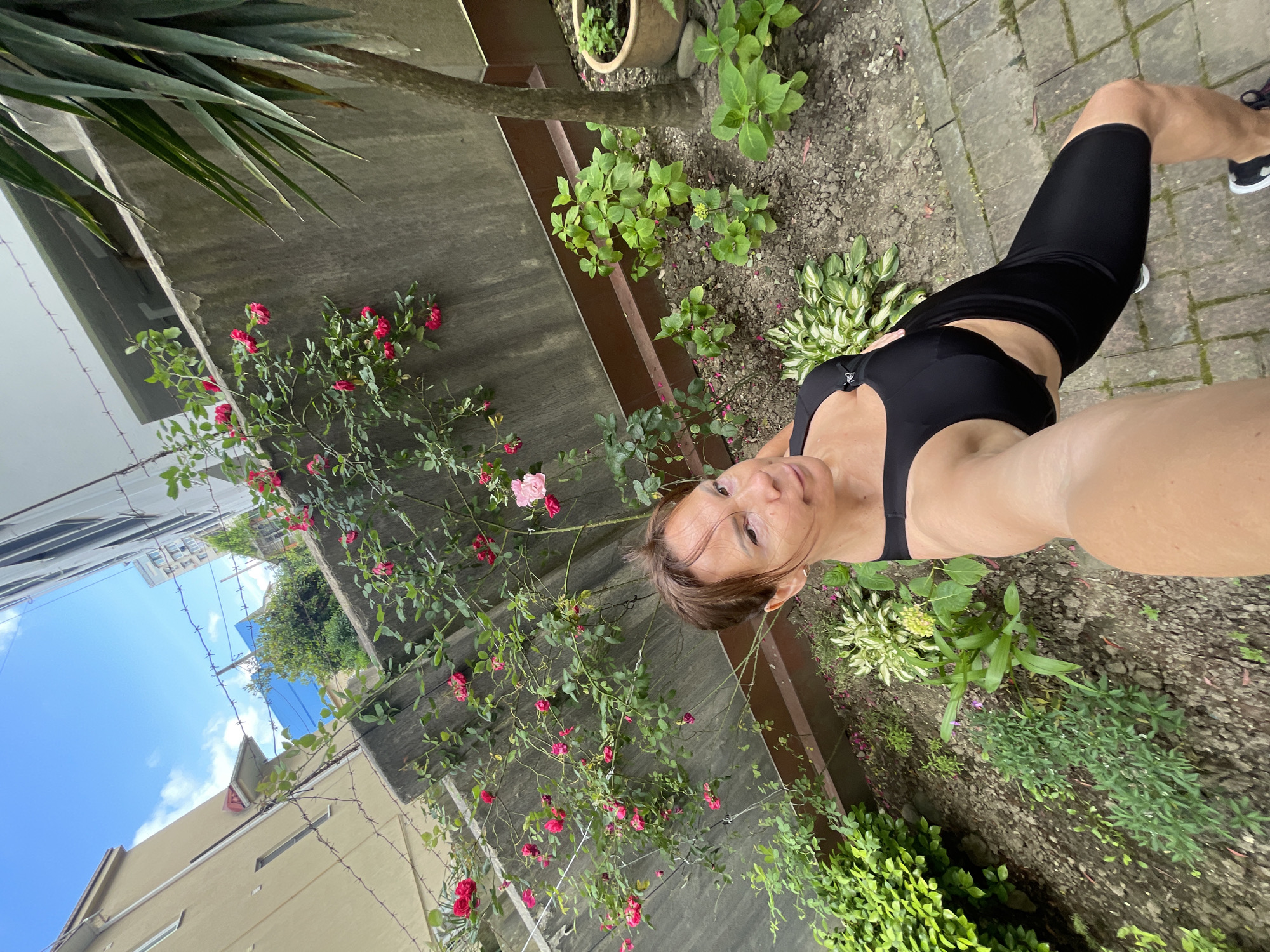 An outdoor, high-angle, selfie-style photograph shows a woman in athletic wear posing in a garden. The woman has brown hair and is wearing a black sports bra and black bike shorts. She is positioned in the lower right portion of the frame. Behind her, a rose bush with pink and red flowers grows along a concrete wall. A palm tree is visible to the right of the rose bush. The garden ground is covered with soil and small plants. In the background, buildings and a blue sky with scattered clouds are visible. The scene appears to be in a residential area.