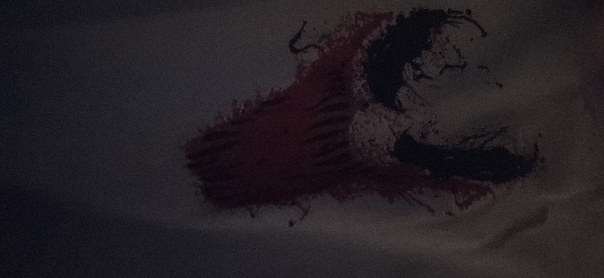 The image displays a dimly lit, abstract design rendered in dark red and black on a lighter, dark grey background, likely fabric or a wall. The dominant feature is a large, textured dark red shape on the left, which appears almost like a stylized, organic mass or a boot-like form. Adjacent to it, on the right, is a fragmented and intricate dark black shape, characterized by irregular edges, splatter patterns, and tendril-like extensions. The interplay of the red and black, along with the jagged and fluid textures, strongly evokes the visual style of the Marvel comic character Carnage, known for his red and black, symbiote appearance. The overall impression is that of a creature or monster, or a dynamic, abstract piece of art. The lighting is very low, making the details subtle. There are no discernible human subjects, explicit location cues for Plovdiv, Bulgaria, or any visible text.