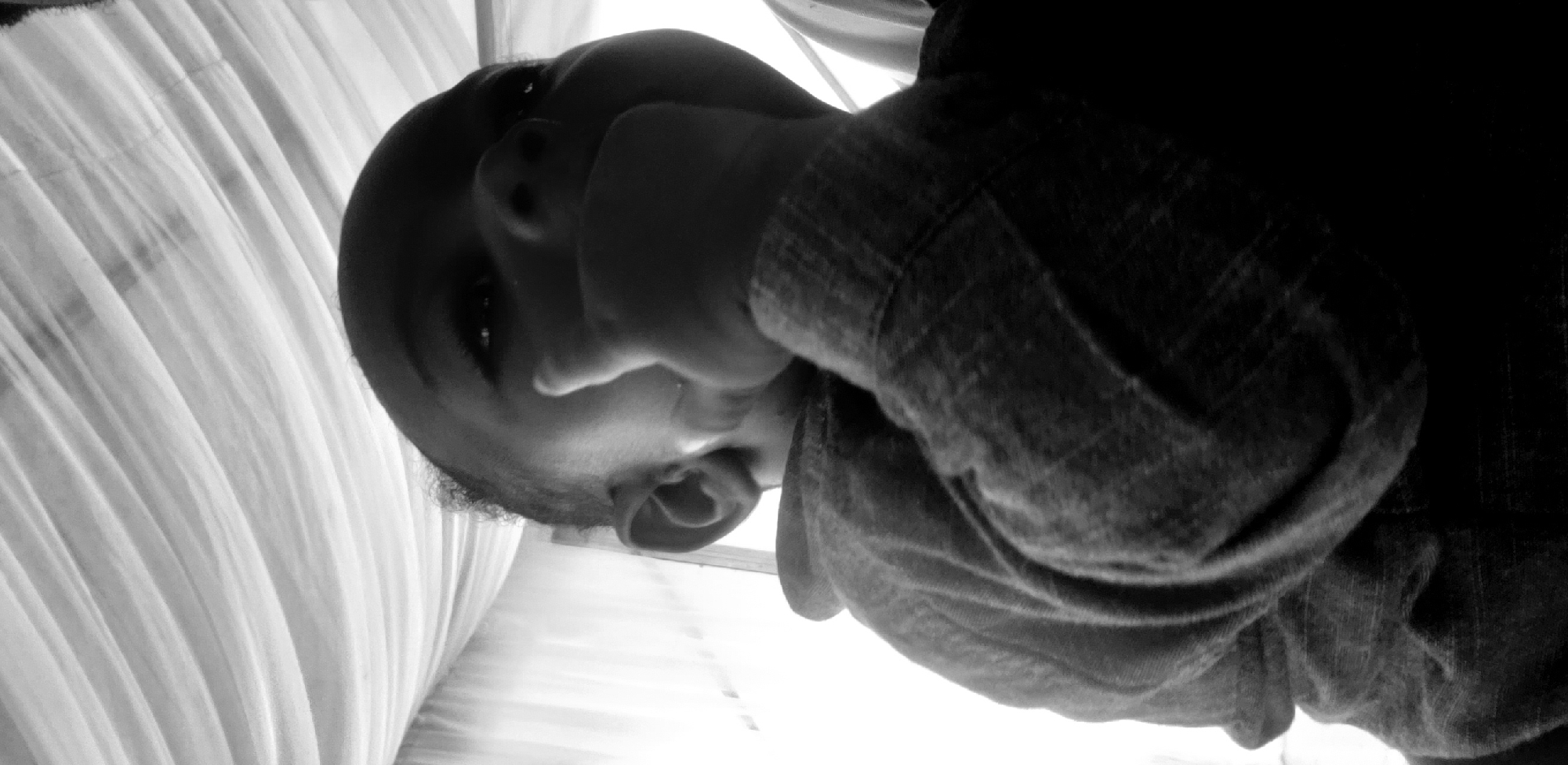 A low-angle, black and white close-up shot captures a young child, their head tilted upwards and to the right, with their right hand held to their mouth. The child appears to be wearing a hooded garment. The background features draped white fabric, possibly curtains or a tent, with light filtering through. The overall impression is of a candid, intimate moment captured in natural light. The image does not contain any discernible urban features or references to a specific location within Narayi, Nigeria.