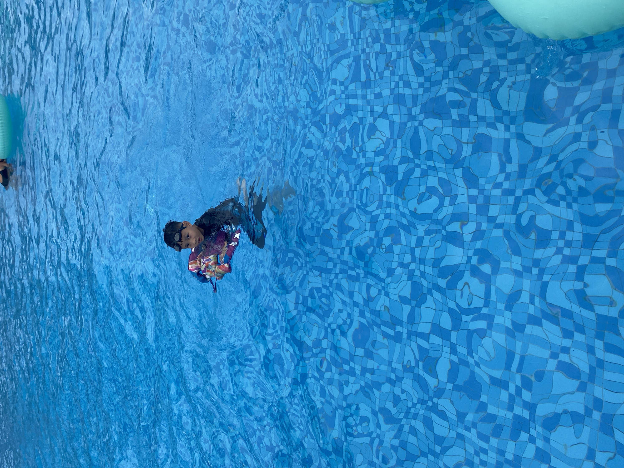 A child wearing goggles and a colorful life vest is swimming in a blue tiled pool. The water surface is rippled. There is also a green inflatable float nearby. The location is Cam Lam, Vietnam.