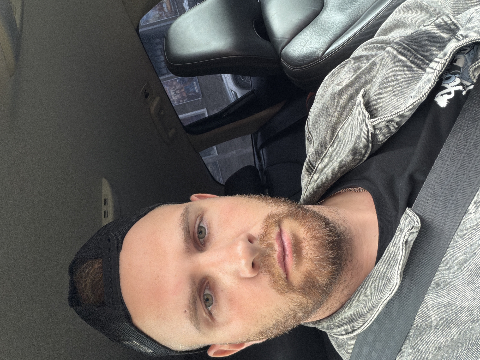 A male individual is seated in the driver's seat of a vehicle, appearing to take a selfie. He has fair skin, a reddish-brown beard and mustache, and greenish-hazel eyes. He wears a black mesh baseball cap with the brim facing forward, a faded grey denim jacket, and a black t-shirt, with a thin gold-colored chain visible around his neck. A black seatbelt is securely fastened across his chest and lap. The car interior features a grey headliner and black leather seats. Through the rear window on the passenger side, portions of a grey building with windows and what appears to be another vehicle (a white car) are visible. Posters featuring multiple figures are affixed to the building's exterior. The scene is located in the city of Votkinsk, Russia.