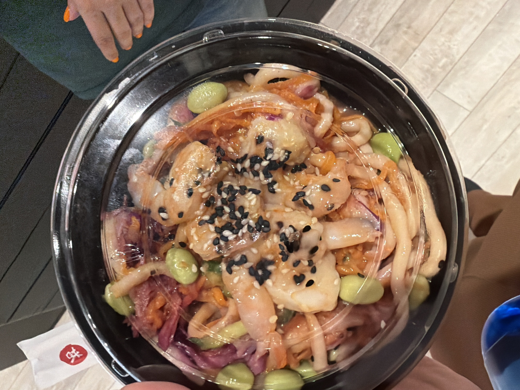 A clear plastic lid covers a black plastic bowl containing a poke bowl. The bowl contains shrimp, edamame, shredded carrots, and red cabbage. The poke bowl is being held by a person's hands. To the left of the bowl, a white packet with a red symbol is visible. In the background, a person's arm with orange nail polish is visible, and the setting appears to be indoors, possibly a food establishment. The flooring is light-colored wood. The scene is illuminated by overhead lighting.
