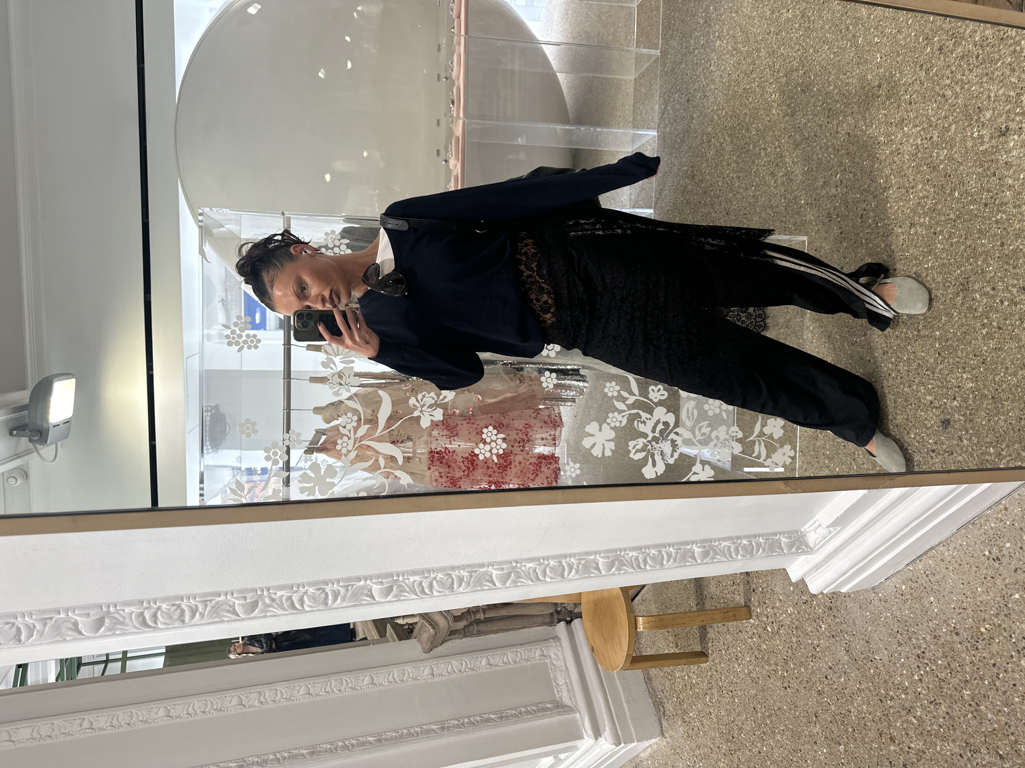 The image is a selfie of a woman in a mirror. She is wearing a dark-colored sweater, a lace skirt, and black pants with white stripes down the side. She has her hair pulled back and is holding a phone in her right hand, taking the photo. The background shows a storefront window with floral decals and display items inside. The floor appears to be terrazzo. The mirror's ornate white frame is visible at the bottom of the image.
While it is difficult to be certain of the specific location without additional information, the architecture and style of the shop interior suggest a location in an upscale commercial area, possibly in a major city like London, United Kingdom.