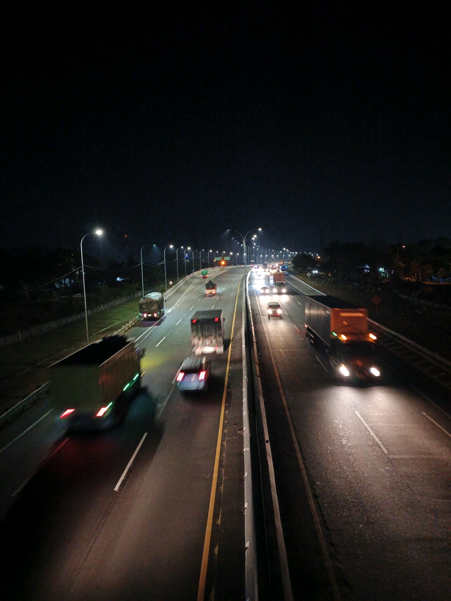 This is a nighttime, eye-level, medium shot of traffic on a multi-lane highway, likely an expressway in or near Kludan, Indonesia. The scene is dark, illuminated by streetlights and the headlights and taillights of the vehicles. The motion blur of the moving vehicles indicates a long exposure photograph.
In the foreground, on the left side of the highway, a large truck with a light-colored cargo bed is moving away from the camera. Its taillights are a streak of red and green. To its right, a silver car is also moving away, with its taillights visible. Further down the lane, another truck is moving away, its taillights blurred red.
In the right lanes, several vehicles are also in motion. A large orange truck is moving towards the camera, its headlights casting a bright glow on the road. To its left, another truck, possibly a box truck, is moving away, its taillights blurred red. Further in the distance, more headlights and taillights indicate a steady flow of traffic. Streetlights line the highway at regular intervals, casting pools of light on the asphalt. The sky is dark and black, with no visible stars or moon. Trees and foliage are visible on the sides of the highway, appearing as dark masses against the night sky. The highway appears to be in good condition, with clear lane markings. The environment suggests an urban or suburban setting on a major transportation route.