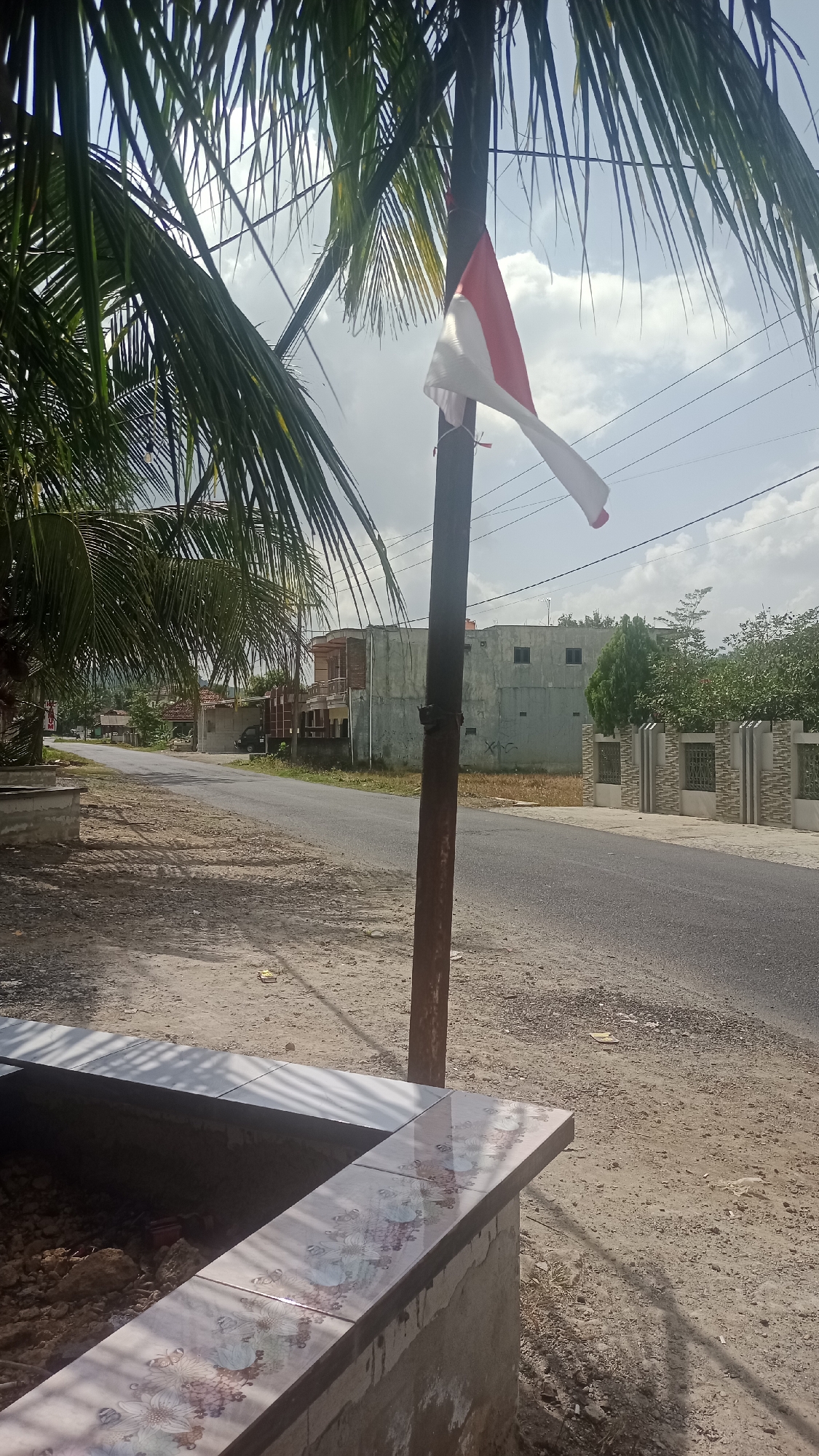 This is a medium-angle, eye-level outdoor photograph taken on a bright, slightly overcast day. The scene depicts a paved road in what appears to be a semi-rural or suburban area of Cibeber, Indonesia. In the foreground, to the left, is a partially constructed concrete structure with a tiled surface featuring a floral pattern. To the right of this structure is a vertical wooden pole with a small Indonesian flag (red and white) attached to it, fluttering in the breeze. Palm fronds from a tree overhang the left side of the frame, casting shadows. The road stretches into the distance, lined with various buildings and some vegetation. On the right side of the road, there is a low decorative wall and some trees. The sky is partly cloudy. There are no people visible in the image.