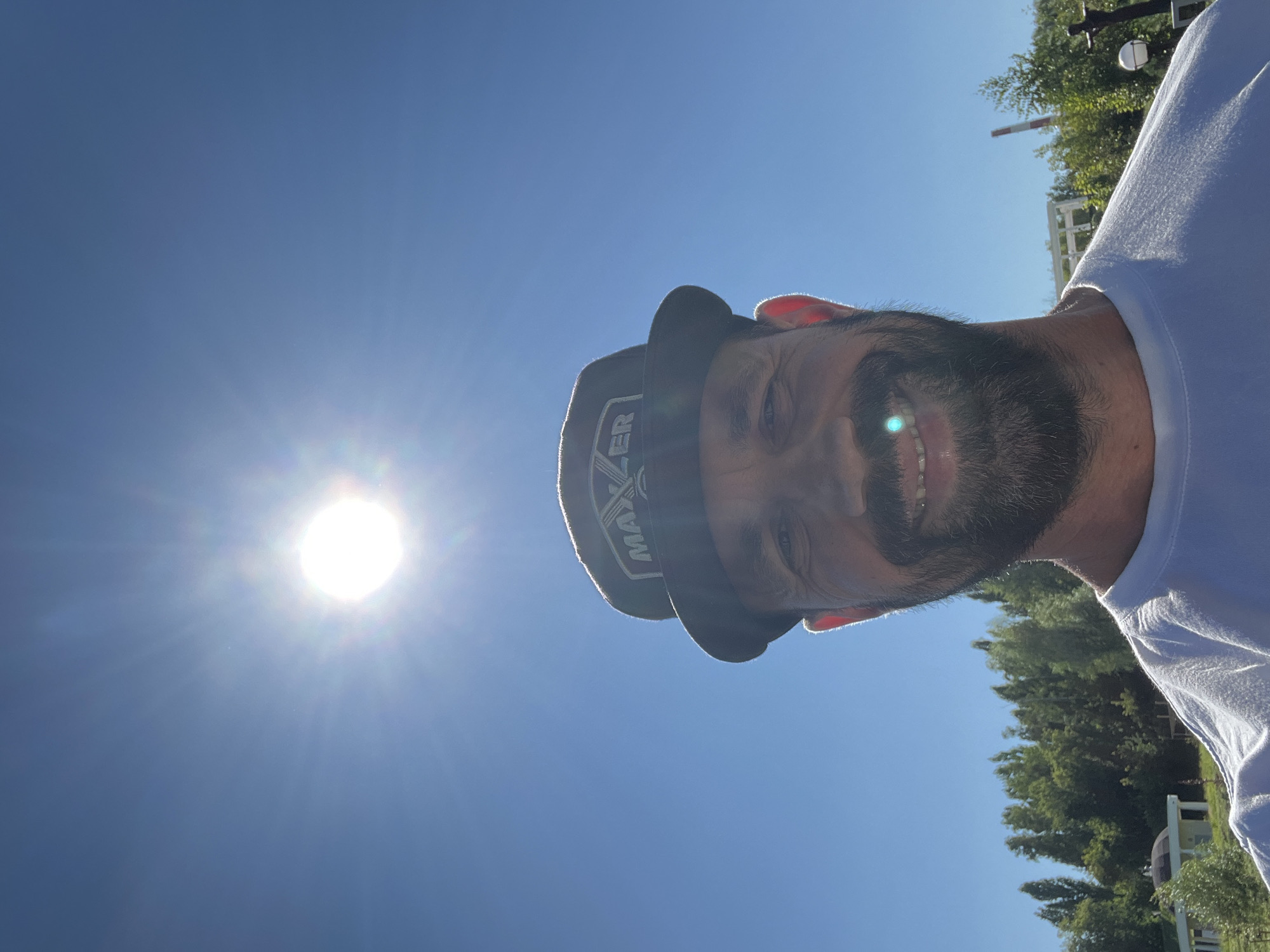 The image is a selfie taken outdoors on a sunny day, with the sun prominently visible in the upper part of the frame. A man is the primary subject, positioned in the lower half of the image. He is wearing a white t-shirt and a dark baseball cap with a logo that appears to say "MAXER". The man has a beard and is smiling, with a bright light reflecting on his teeth. The background shows a clear blue sky, a line of trees, and some distant structures, including what appears to be a building with a domed roof and a factory chimney. The lighting suggests it is daytime. Based on publicly available information, the presence of specific industrial structures and the general landscape is consistent with areas within the city of Cheboksary, Russia.