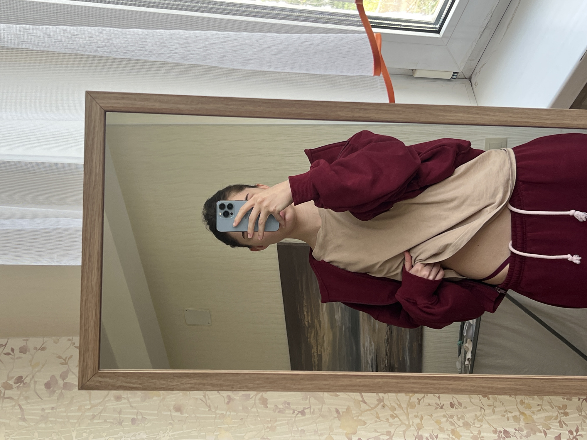 An individual is captured taking a mirror selfie, reflected with their head on the left and body extending to the right, suggesting the image is rotated 90 degrees counter-clockwise. The person, appearing young, has dark hair and is holding a light blue/grey smartphone with multiple camera lenses to take the picture. They are wearing a maroon hoodie or sweatshirt, and a light beige t-shirt which is lifted to reveal part of their abdomen and a dark bralette or bikini top. Matching maroon sweatpants with a white drawstring are also visible.
The mirror has a simple rectangular shape with a light brown wooden frame. The actual background of the room features a window with white horizontal blinds at the top, allowing natural daylight to filter in and revealing a hint of greenery outside. An orange strap hangs near the window. The wall below the mirror is adorned with wallpaper featuring subtle, light-colored floral or leafy patterns, possibly in shades of pale pink, purple, and beige, against a cream background.
Reflected in the mirror, behind the person, is a plain beige wall, a white rectangular wall-mounted object (possibly an outlet or switch), and a large, abstract painting with horizontal strokes of dark brown, grey, and cream. Hints of other room elements or furniture are also faintly visible in the reflection. The overall lighting suggests it is daytime. The setting, located in Saint Petersburg, Russia, appears to be an indoor living space. There is no visible text.