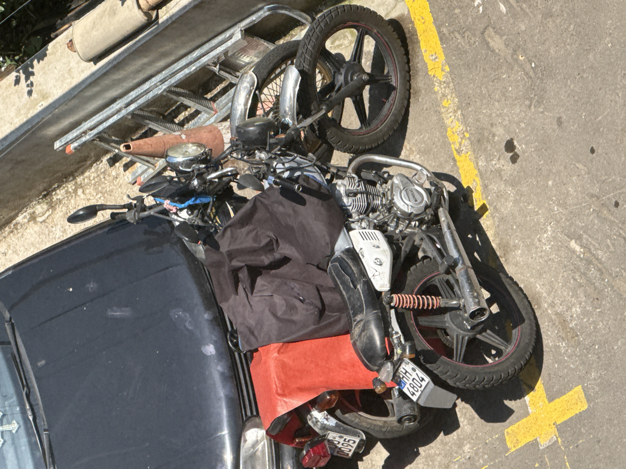 An eye-level shot captures a parked motorcycle in what appears to be a city environment. The motorcycle, a gray and black model with a red covering over its seat, is positioned in front of a black car. The motorcycle's license plate displays the characters "HH 4804". In the background, a ladder and a traffic cone are visible, suggesting an area undergoing construction or maintenance. The ground is asphalt with yellow markings, indicative of a parking lot or roadway. Sunlight casts shadows, indicating it is daytime. The scene appears to be in Tbilisi, Georgia, based on the style of the license plate and the general urban aesthetic. There are no people visible in the image.