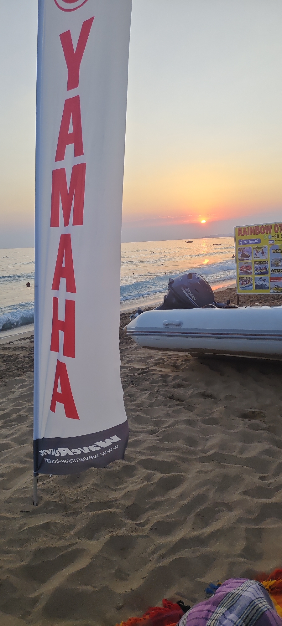 The image shows a beach scene in Alanya, Turkey, at sunset. A large white banner with red "YAMAHA" lettering is prominent in the foreground, positioned vertically. Behind the banner, a white inflatable boat with a Yamaha outboard motor is parked on the sandy beach. The sea is visible with gentle waves and a few people swimming or wading in the water. In the distance, the sun is setting over the horizon, casting a warm glow on the sky and water. A yellow advertising sign is also visible, promoting "RAINBOW 07" activities with images of jet skis and parasailing. The sand is rippled, suggesting it's a well-used beach. A colorful fabric, possibly a towel or sarong, is partially visible in the lower right corner.