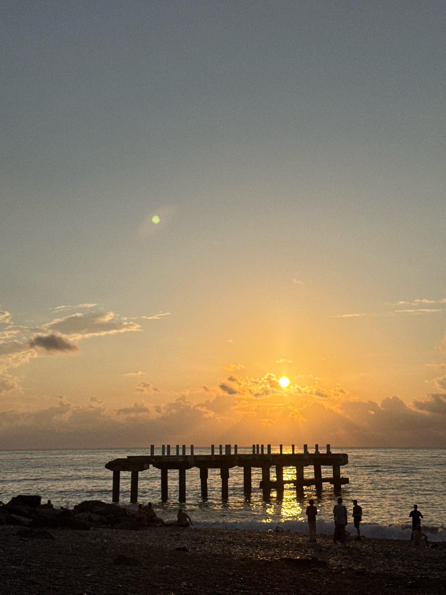 The image depicts a sunset scene at a beach in Batumi, Georgia. In the foreground, a pebbled beach is visible, with several people scattered across it, some appearing to be sitting and others standing. To the left, there is a pile of large rocks. In the middle ground, the sea is calm, reflecting the warm hues of the sunset. An old, partially damaged pier extends into the water, supported by concrete pillars. The pier's structure is rudimentary, with vertical posts along its edge. In the background, the sun is low on the horizon, casting a golden glow on the water and illuminating the clouds. The sky transitions from a pale blue at the top to orange and yellow near the horizon, with scattered clouds adding texture to the scene.