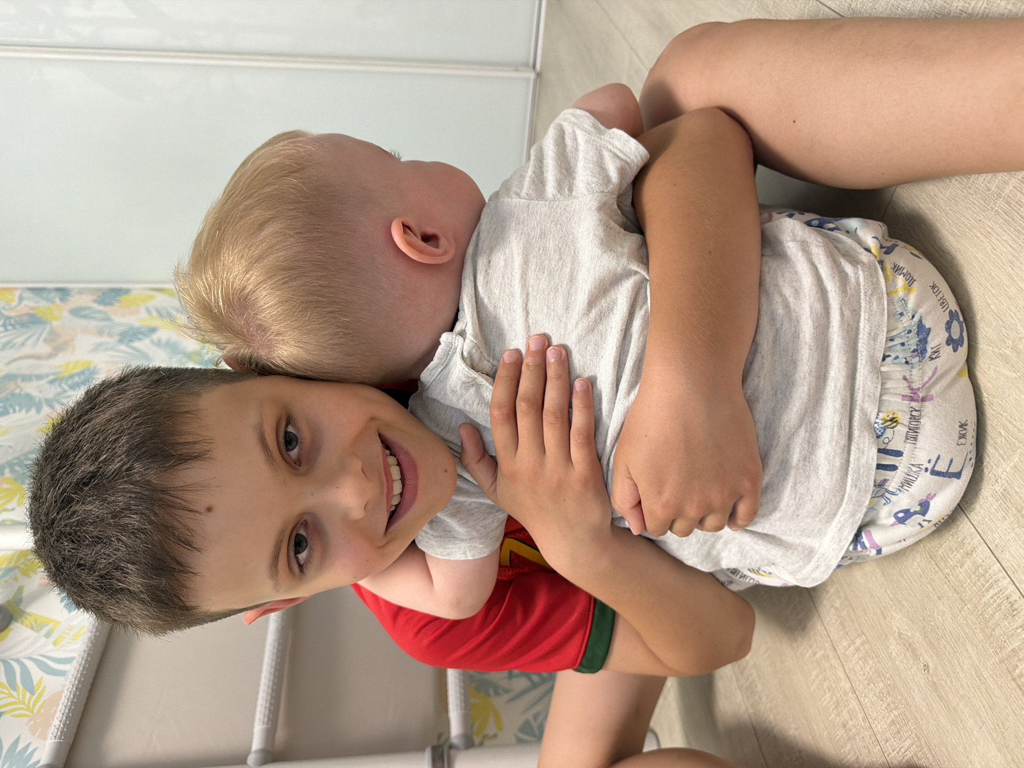 The image captures a heartwarming indoor moment between two boys, likely siblings, in what appears to be a home in Votkinsk, Russia. The scene is brightly lit, suggesting daytime.
The older boy, with short dark hair and dark eyes, is positioned in the foreground, his face turned towards the camera with a wide, joyful smile that reveals his teeth. He is wearing a red t-shirt with a green stripe at the bottom of the sleeve.
Embracing him from behind is a younger boy, a toddler with short, light blonde hair. His face is nestled against the older boy's head and shoulder, with his ear visible, and his posture suggests comfort and affection. He wears a light grey short-sleeved t-shirt. On his patterned pull-up diaper or underwear, several Russian words are visible, including "ЛИСТОК" (leaf), "ДОМИК" (little house), "МЫШКА" (mouse), "ЦВЕТОК" (flower), "ВОТКИНСК" (Votkinsk), and "ПЯТИГОРСК" (Pyatigorsk), alongside cartoon-like illustrations of animals and objects.
The background features light wooden floorboards and a light-colored wall. On the left side, a section of wallpaper or fabric with a delicate pattern of light blue, yellow, and green leaves or floral motifs is visible. The overall impression is one of tenderness and brotherly love in a domestic setting.