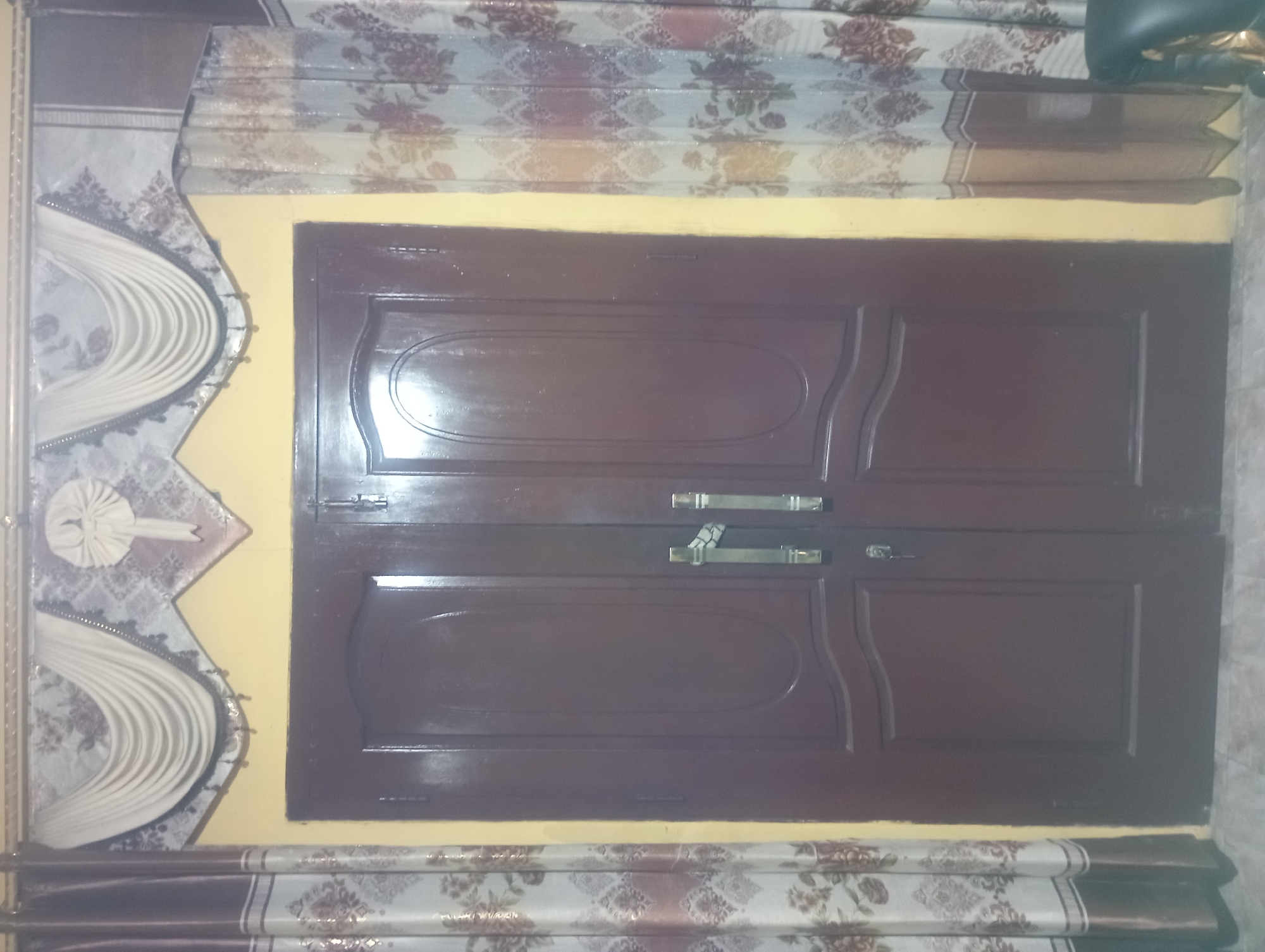 A brown double door is centered in the frame, flanked by floral curtains. Above the door, a decorative valance with draped fabric and beaded trim hangs across the top of the doorway. The wall behind the door and curtains is painted yellow. The floor appears to be tiled. To the right of the door, the corner of a dark, upholstered chair is visible. The scene suggests an interior setting, possibly a home in Maos Kidul, Indonesia, given the context.