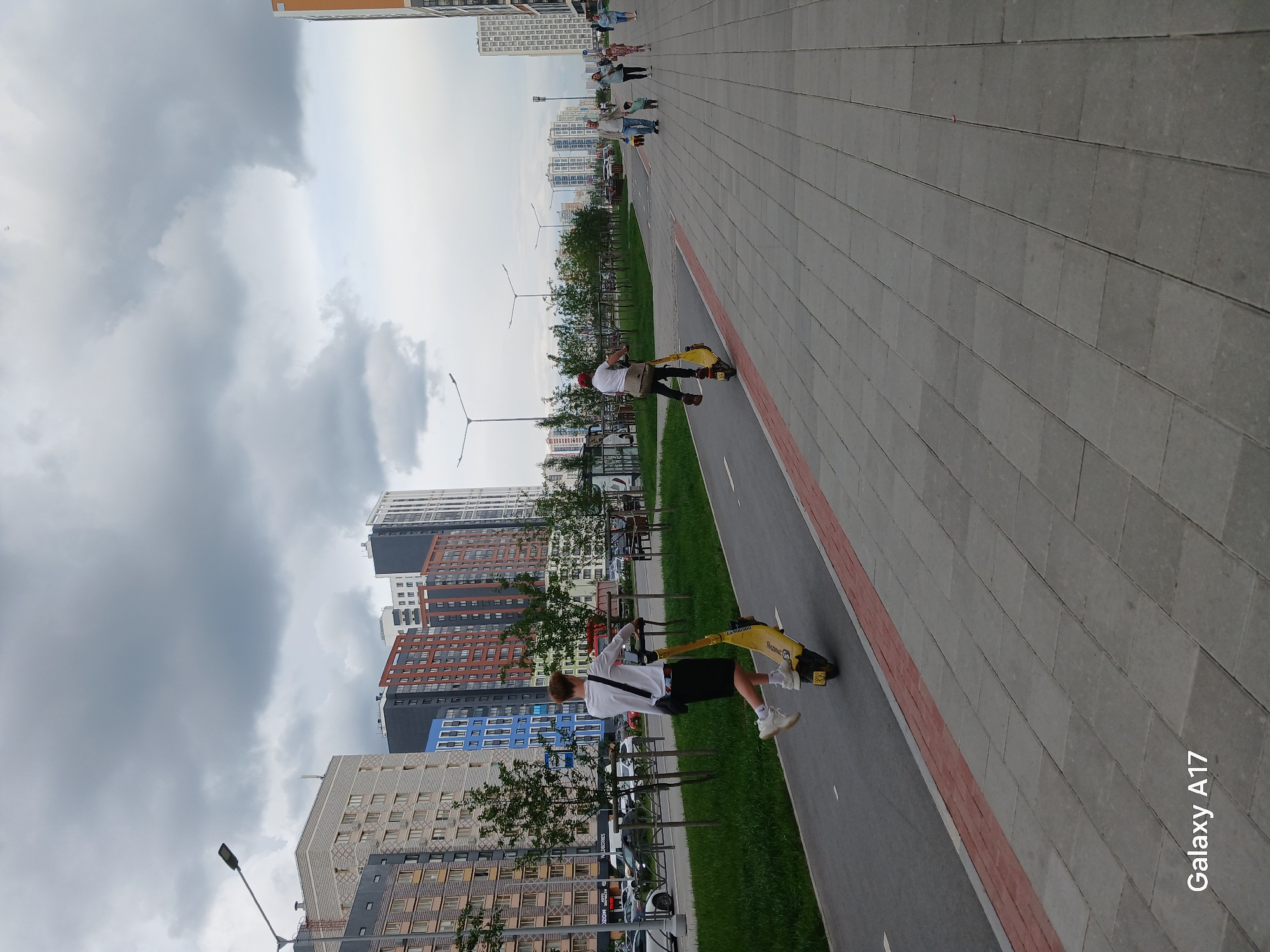 In Yekaterinburg, Russia, on a cloudy day, two individuals are riding electric scooters along a paved pathway that runs parallel to a road. The pathway is separated from the road by a strip of green grass and trees. Beyond the road, a line of multi-story residential buildings with diverse architectural styles and color schemes is visible. Several vehicles are parked along the road. A group of people, including adults and children, are walking on the sidewalk on the right side of the image. The sky is overcast with heavy clouds, suggesting the possibility of rain. The overall scene depicts an urban environment with recreational activity taking place in a public space.