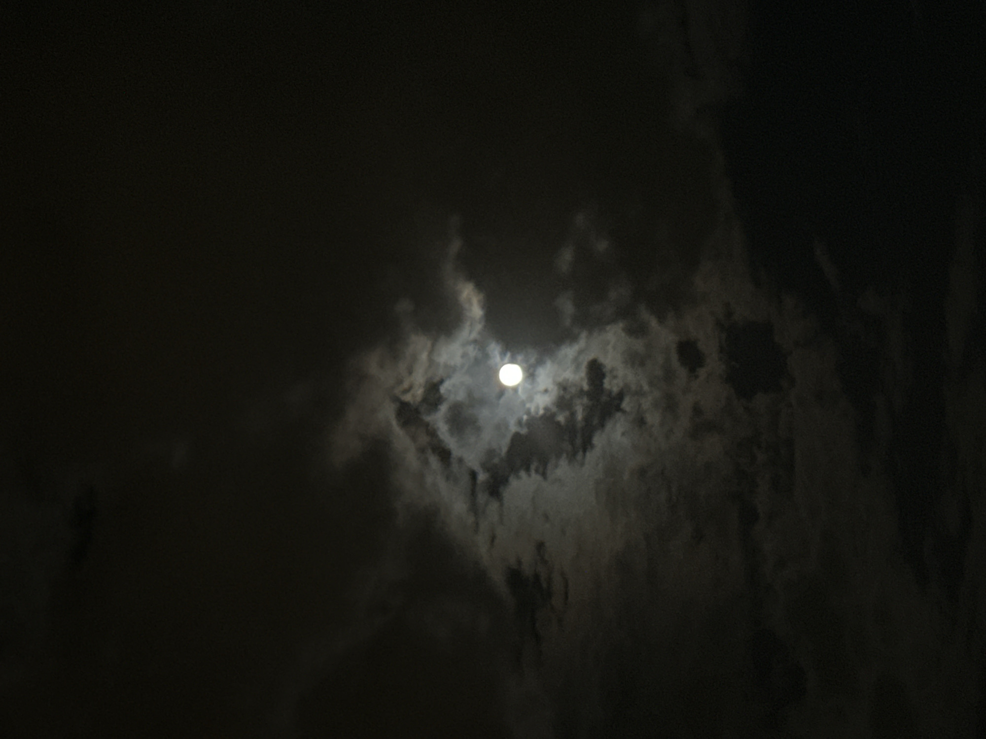 The image presents a dark, nocturnal sky with the moon partially obscured by clouds. The central feature is the bright, circular moon, which serves as a powerful light source against the deep black of the night. Numerous clouds are visible, characterized by varying densities and forms. The moonlight illuminates the clouds from behind and within, creating distinct contrasts; the areas directly hit by the moon's light appear brilliant white or light grey, while other parts remain darker, rendered in shades of grey and black.
The scene captures a moment of natural atmospheric interaction between the moon and clouds at night. There are no human subjects, man-made objects, or discernible terrestrial landscapes, focusing entirely on the celestial display. The time of day is clearly night, and the weather condition is cloudy. No specific location cues are visible within the image to tie it to Yekaterinburg, Russia, beyond the provided context. There is no visible text. The overall impression is one of serene darkness punctuated by the ethereal glow of the moon filtering through the clouds.