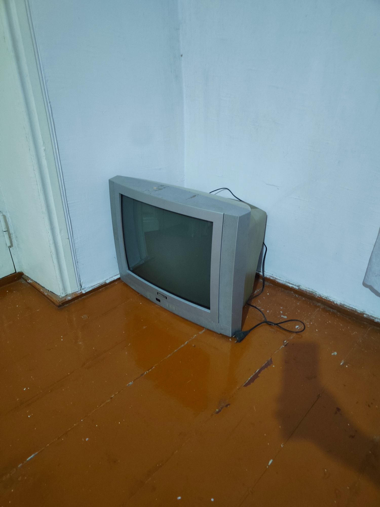 An old, grey, CRT television set is positioned in the corner of an empty room. The television, which appears to be turned off, rests on a wooden floor that is a deep reddish-brown color and shows signs of wear, with visible planks, scratches, and scattered white debris. The walls are painted a light, neutral color, and a white doorframe is visible to the left of the television, extending from the floor upwards and meeting the wall at a right angle. A black power cord snakes from the back of the television across the floor, ending in a plug that is not connected to an outlet. The lighting in the room casts a long shadow from the television onto the floor.