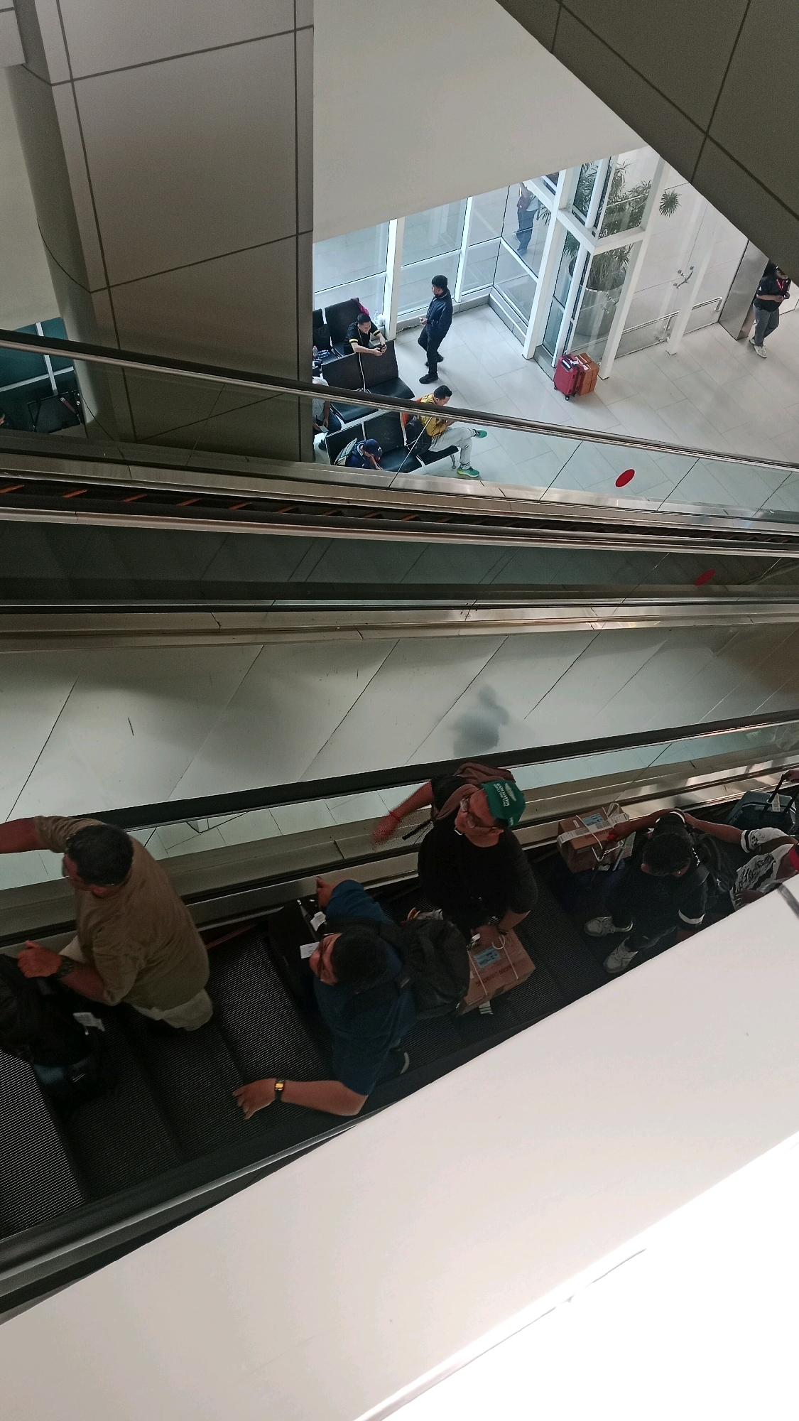 The image shows an interior scene, likely an airport or a large modern building, with an escalator in the foreground. Several people are visible on the escalator and in the background.
In the foreground, on the escalator moving downwards, there are at least three individuals. The person closest to the bottom of the frame is wearing a light brown long-sleeved shirt and appears to be holding onto the handrail. To their right and slightly further up the escalator, a person wearing a dark blue shirt with a backpack is visible, also holding the handrail. Another person to their right, wearing a dark t-shirt and a green cap, is also on the escalator, appearing to reach out towards the handrail. Luggage is present on the escalator near some of these individuals.
In the background, on a different level, there is a waiting area with seating. Several people are seated and standing. One person in a dark jacket is standing near a large window. Another person in a yellow shirt is seated. A red suitcase is visible on the floor in the background. The architecture suggests a contemporary design with large windows and metallic accents.
The scene depicts a common travel or transit environment. The presence of escalators, seating areas, and luggage indicates a location where people are moving between different levels or waiting for transport. The overall impression is one of a busy, functional space. The mention of Tangerang, Indonesia, suggests this location is within that city.
