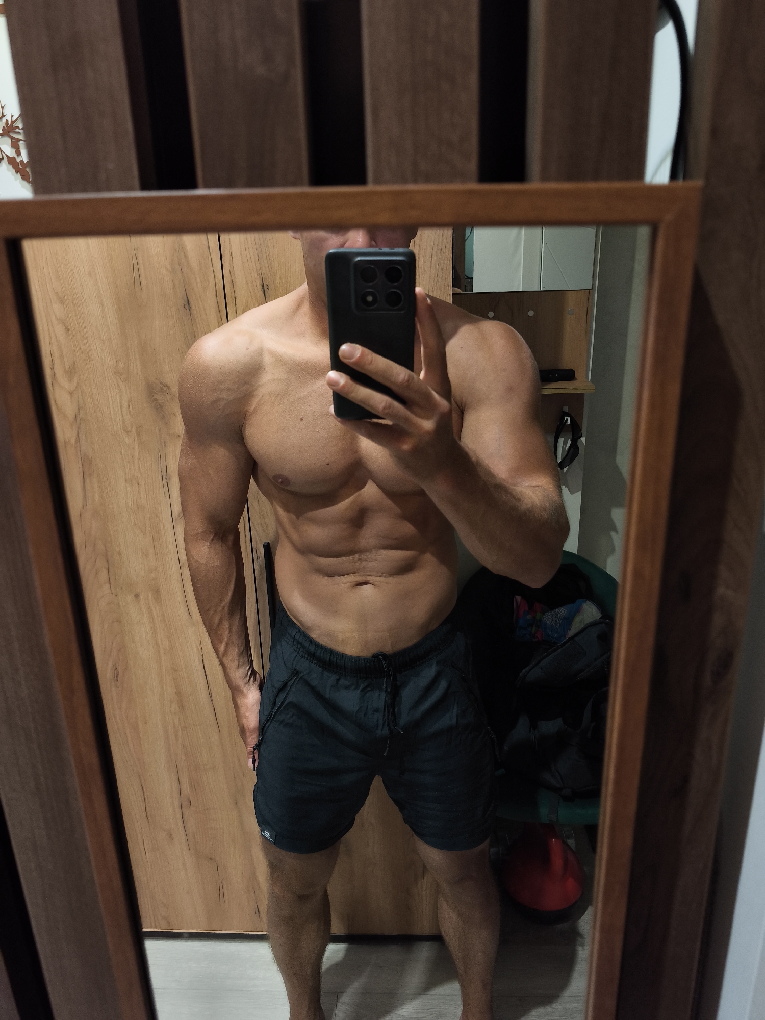 A man with a muscular physique stands in front of a mirror, taking a selfie with his phone. He is wearing dark shorts. The background shows a wooden wardrobe and a glimpse of other items in the room. The scene appears to be indoors.
