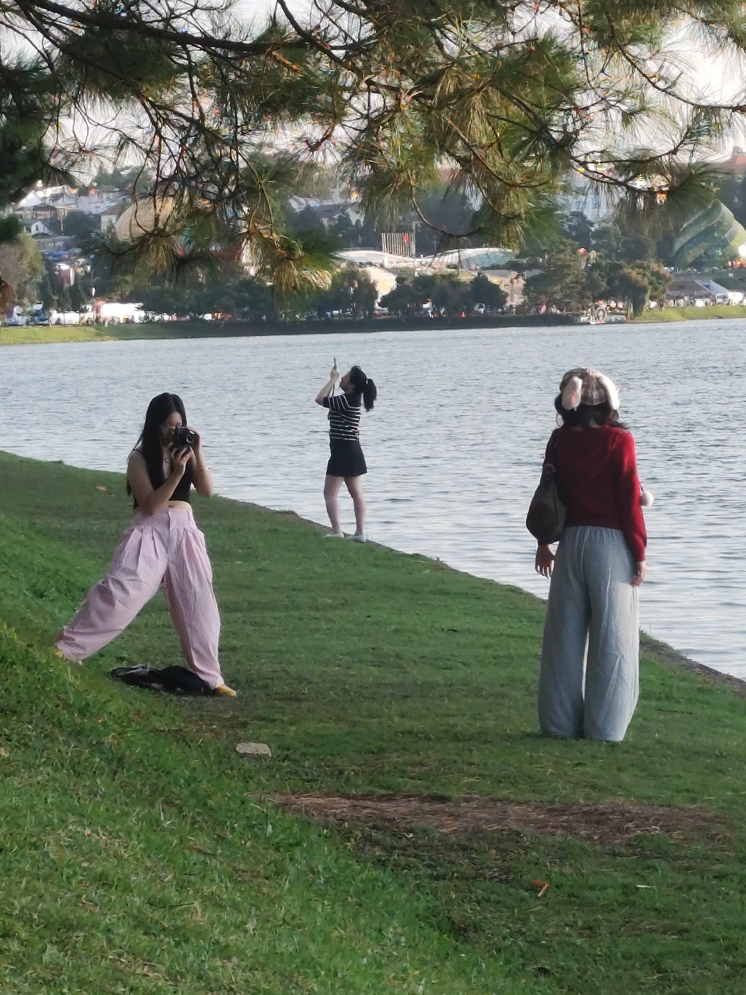The image shows a scenic view of Xuan Huong Lake in Da Lat, Vietnam, with pine trees in the foreground. Three individuals are visible on the grassy shore. On the left, a woman in pink wide-leg pants and a black top is taking a photo with a camera. In the middle, another woman in a striped shirt and black skirt is holding up her phone, possibly taking a picture or video. On the right, a third person, facing away from the camera, is wearing a red hoodie, light gray wide-leg pants, and a hat with ear flaps, carrying a dark bag over their shoulder. The lake is calm, and the background reveals a cityscape with buildings and some greenery.