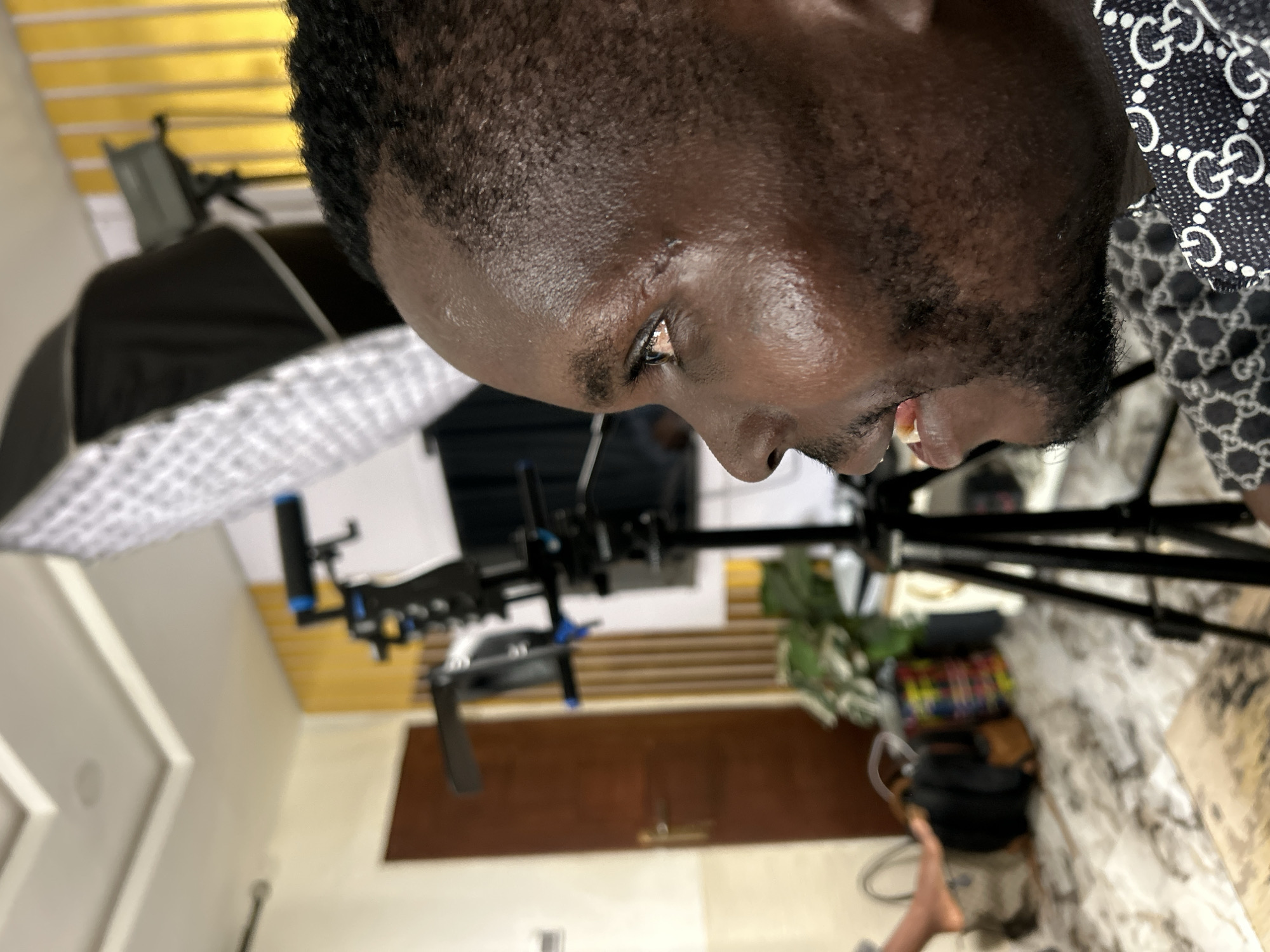 A medium shot, taken at a severe angle, features a dark-skinned male subject, from the nose up, positioned in the upper right corner. The subject has close-cropped hair and a trimmed beard. He is wearing a patterned black and white garment near his ear.
In the midground, a camera rig on a stand is centrally positioned. An octagonal softbox, partly obscured by the camera rig, is in the background on the left. A partially obscured wooden door is behind the camera rig, slightly off-center. A multi-colored item rests on the floor to the right of the camera. The setting appears to be an interior location.
The city location is Unknown, Unknown.