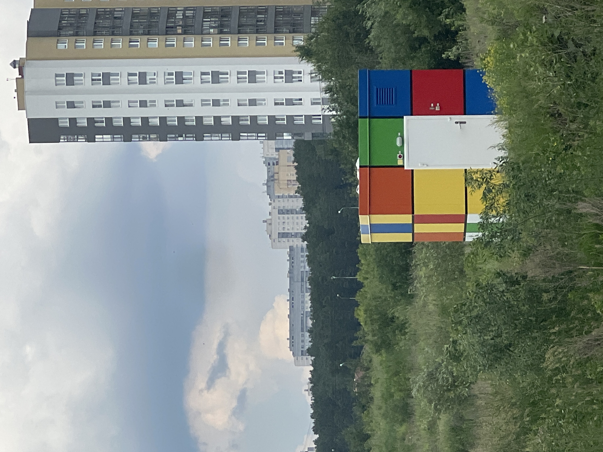 The image shows a colorful, cube-shaped structure resembling a Rubik's Cube, located in a grassy, overgrown area. The structure is divided into colored panels, with a white door on the front. In the background, a tall, modern apartment building with a gray, white, and yellow facade stands against a cloudy sky. Further in the distance, other buildings and a dense line of trees are visible. The overall scene suggests an urban or suburban setting. This structure is likely a utility box or shelter. The style of the apartment building, with its modern design and color palette, is common in new residential developments in Russian cities. The presence of such structures in Yekaterinburg, Russia, is consistent with urban development trends.