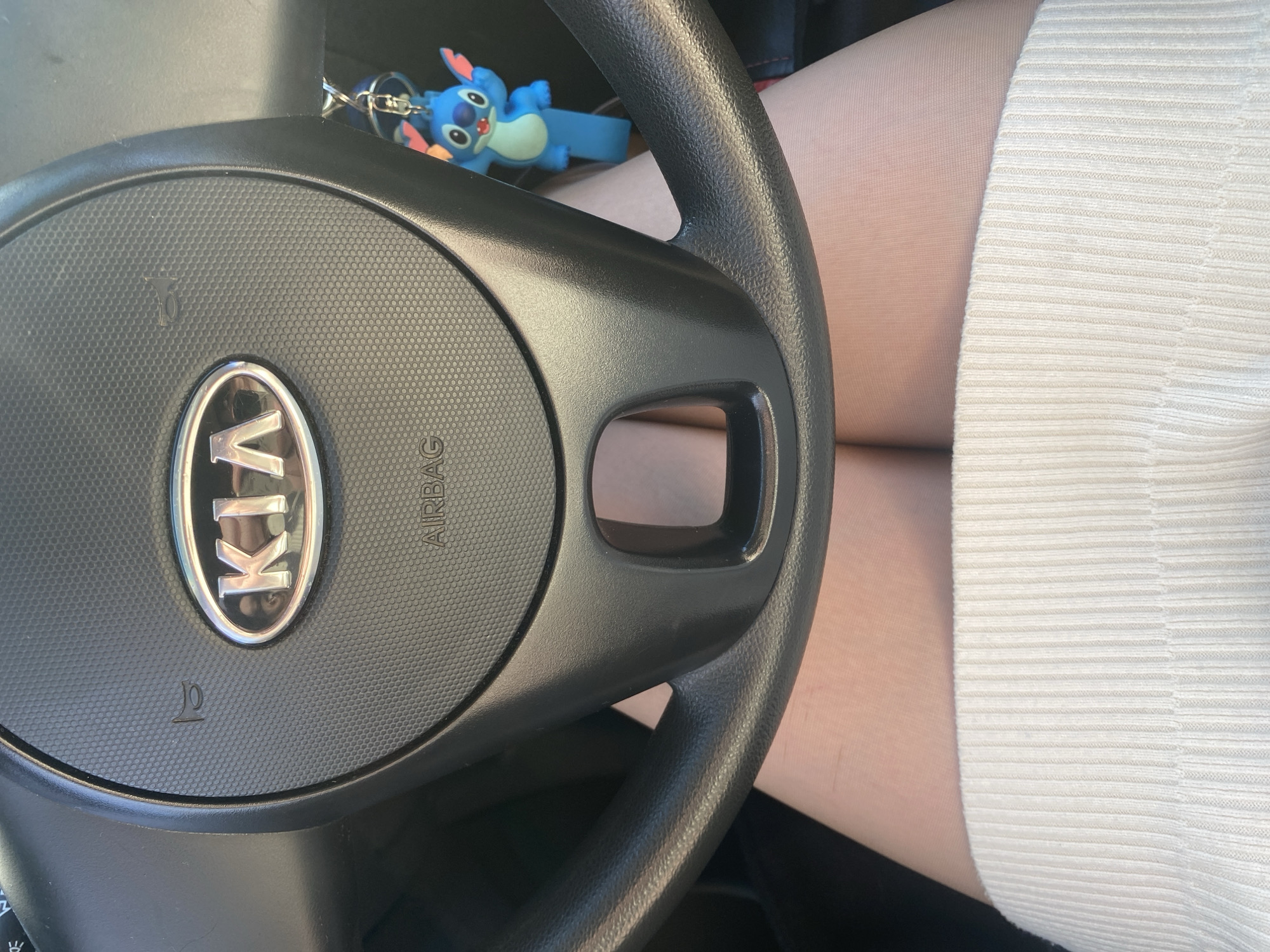 A photograph shows the steering wheel of a Kia vehicle. A blue Stitch keychain is attached to the steering wheel. A person wearing pale pantyhose and a ribbed white skirt is positioned behind the steering wheel. The setting is inside a vehicle, but the location in Chaikovsky, Russia, cannot be determined from the image.