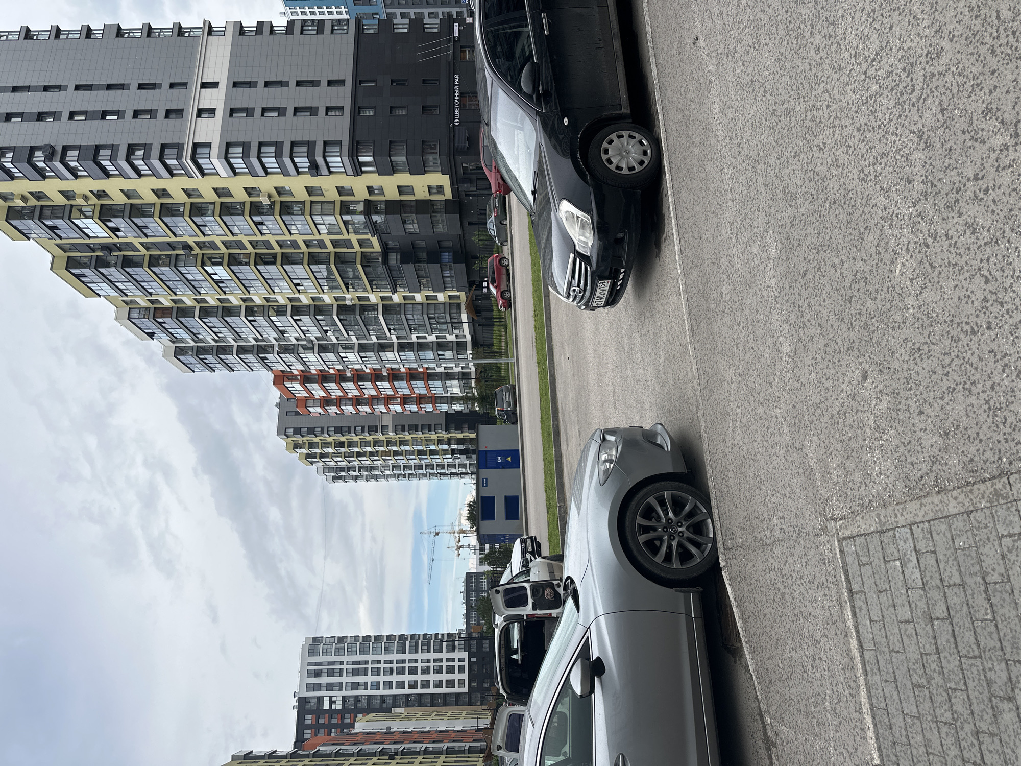The image captures a street scene in Perm, Russia, characterized by modern high-rise residential buildings and parked vehicles under an overcast sky. The photo appears to be taken from ground level, looking towards the buildings.
In the foreground and mid-ground, an asphalt road or parking area stretches across the frame. Two cars are prominently parked along the right side of the road. Closest to the viewer is a silver/grey sedan (possibly a Mazda), showing its front and right side. Further down the road is a black crossover SUV, displaying its front and right side, with the Russian license plate "O740TH 159" clearly visible, confirming the Perm region. In the background, several other parked vehicles can be seen, including a red car and a white commercial van with its rear doors open.
The left and background sections of the image are dominated by numerous tall, contemporary apartment buildings. These buildings feature diverse architectural styles and color palettes, including facades of grey, black, yellow, orange, and white, with many large windows and balconies. On one of the black and yellow buildings, the text "(ЦВЕТОЧНЫЙ РАЙ)" which translates to "Flower Paradise," is visible. In the distant background, beyond the immediate residential structures, construction cranes indicate ongoing development in the area. A small blue container or building marked "B4" is also present in the mid-ground.
The sky is largely cloudy, suggesting an overcast day. No people are visible in the scene. The overall impression is that of a contemporary urban residential district, likely a newly developed area, with cars parked along a street.